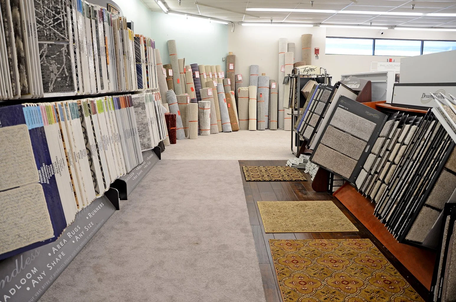 Carpet and rug samples displayed on racks and stands in a flooring store, with rolled rugs stacked in the background.