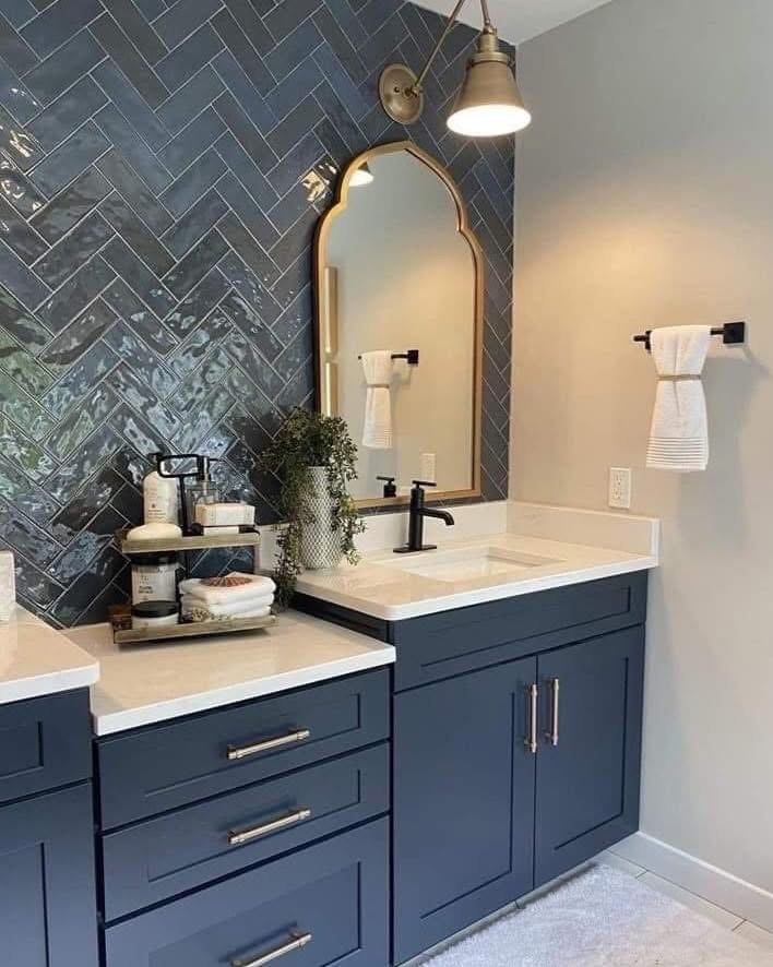 Bathroom vanity with navy blue cabinets, white countertop, black faucet, oval mirror with a gold frame, gold wall sconce, towel rack with a white towel, decorative plant, and various toiletries on a tray.