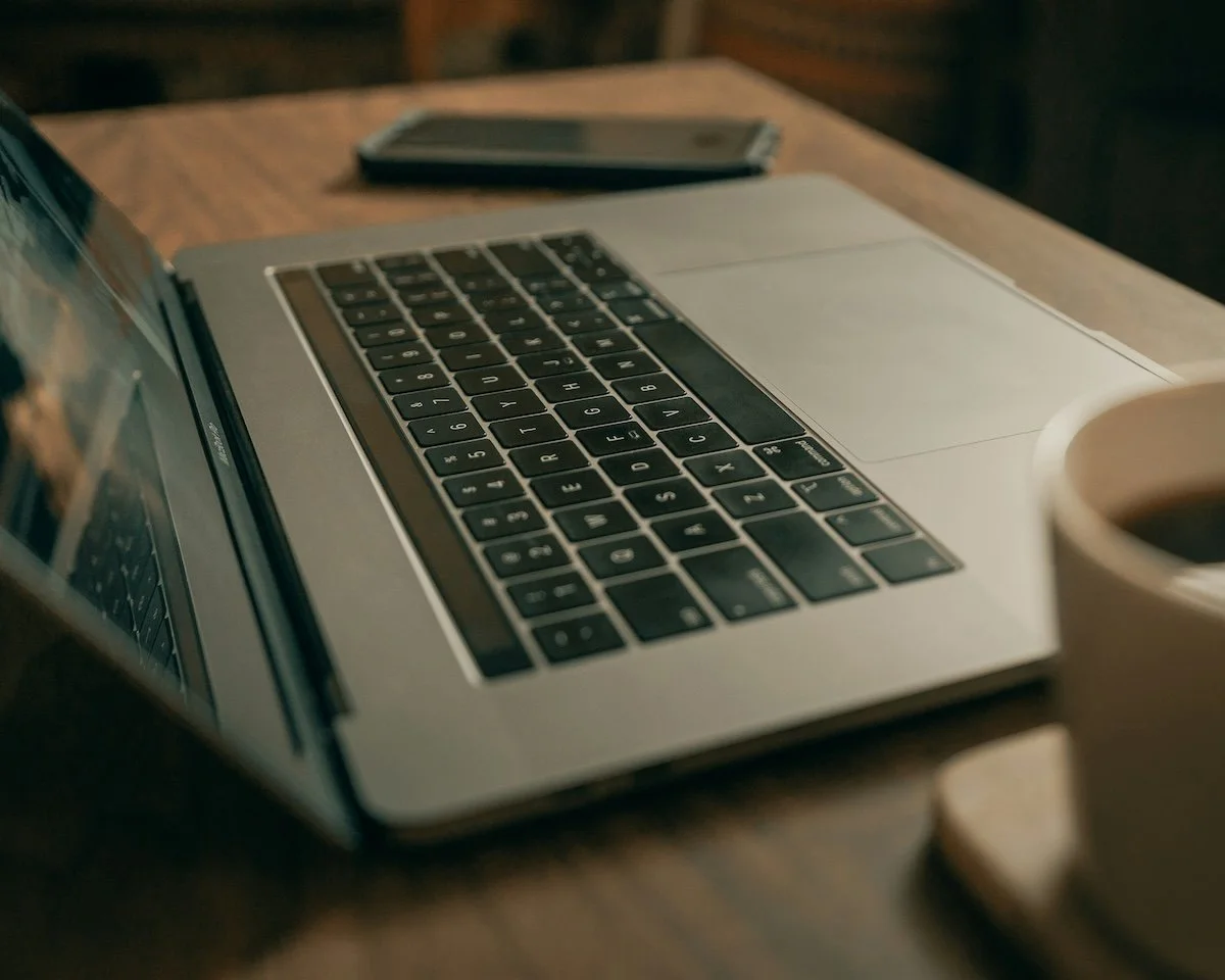 A silver laptop computer on a wooden table, alongside a smartphone and a cup of coffee.