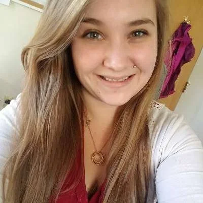 Young woman with long blonde hair taking a selfie indoors, smiling at the camera.
