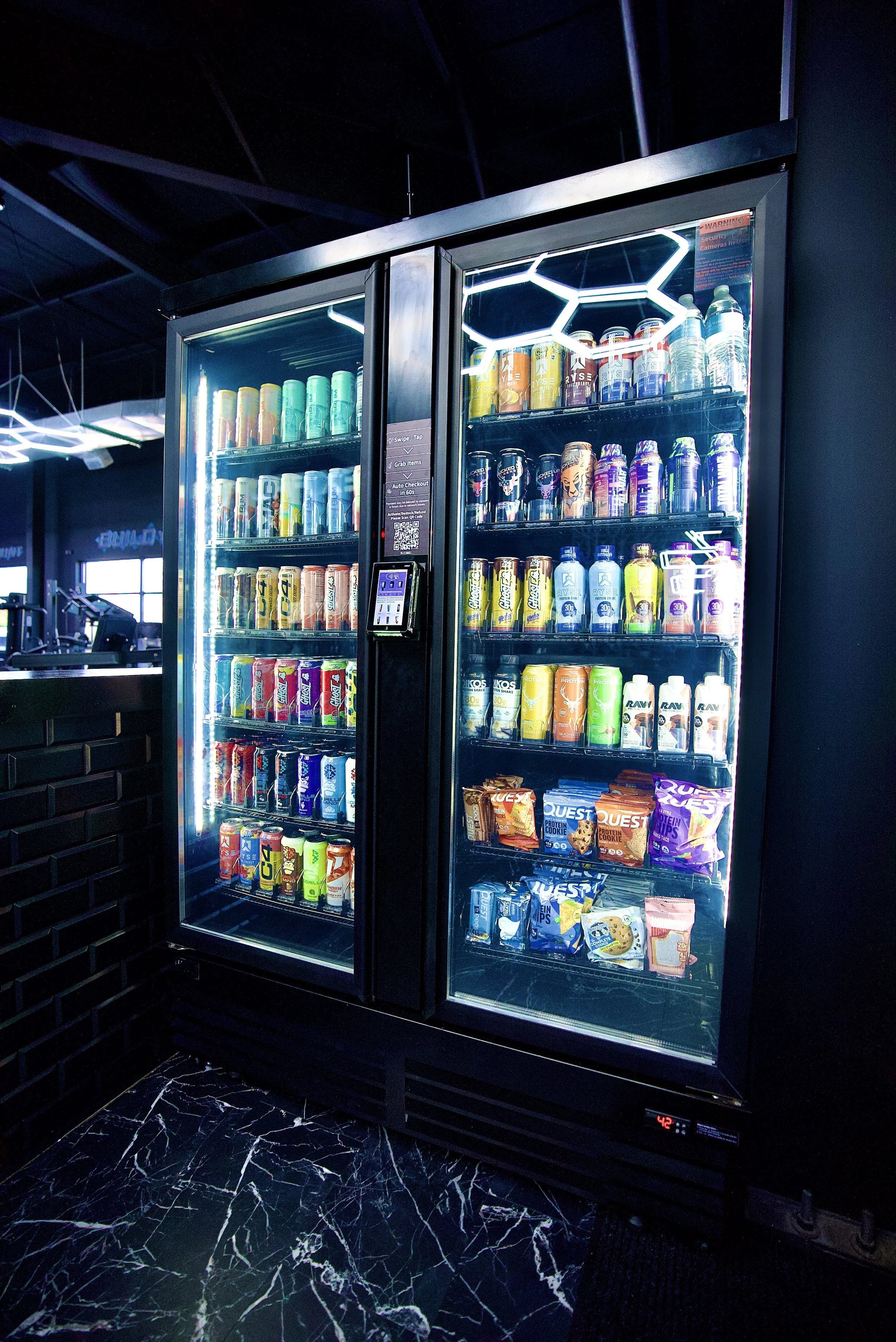 A vending machine labeled '24H SMART VENDING' stocked with energy drinks and canned beverages inside a gym or fitness center.