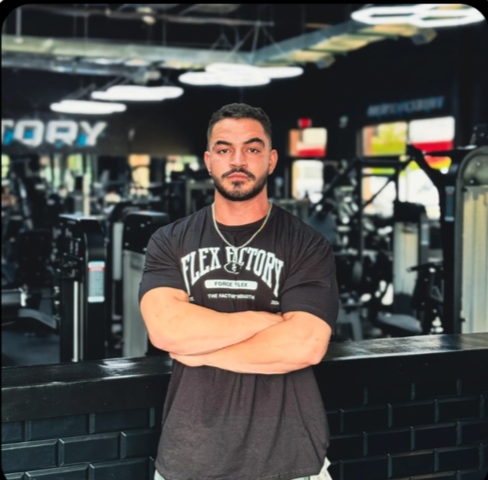 A man with short dark hair and a beard standing with crossed arms in a gym, surrounded by exercise equipment.