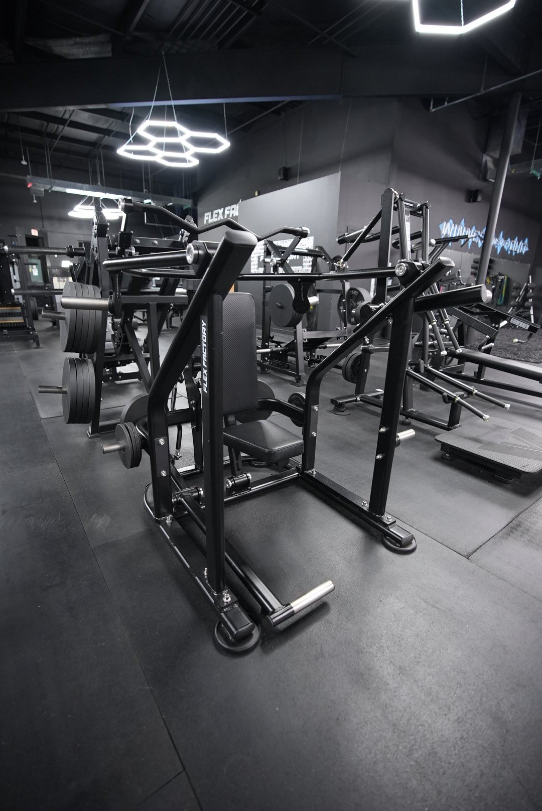 Inside a modern gym with various black fitness equipment, including a leg press machine, weight racks, and a treadmill, with a dark ceiling and illuminated hexagon-shaped ceiling lights.