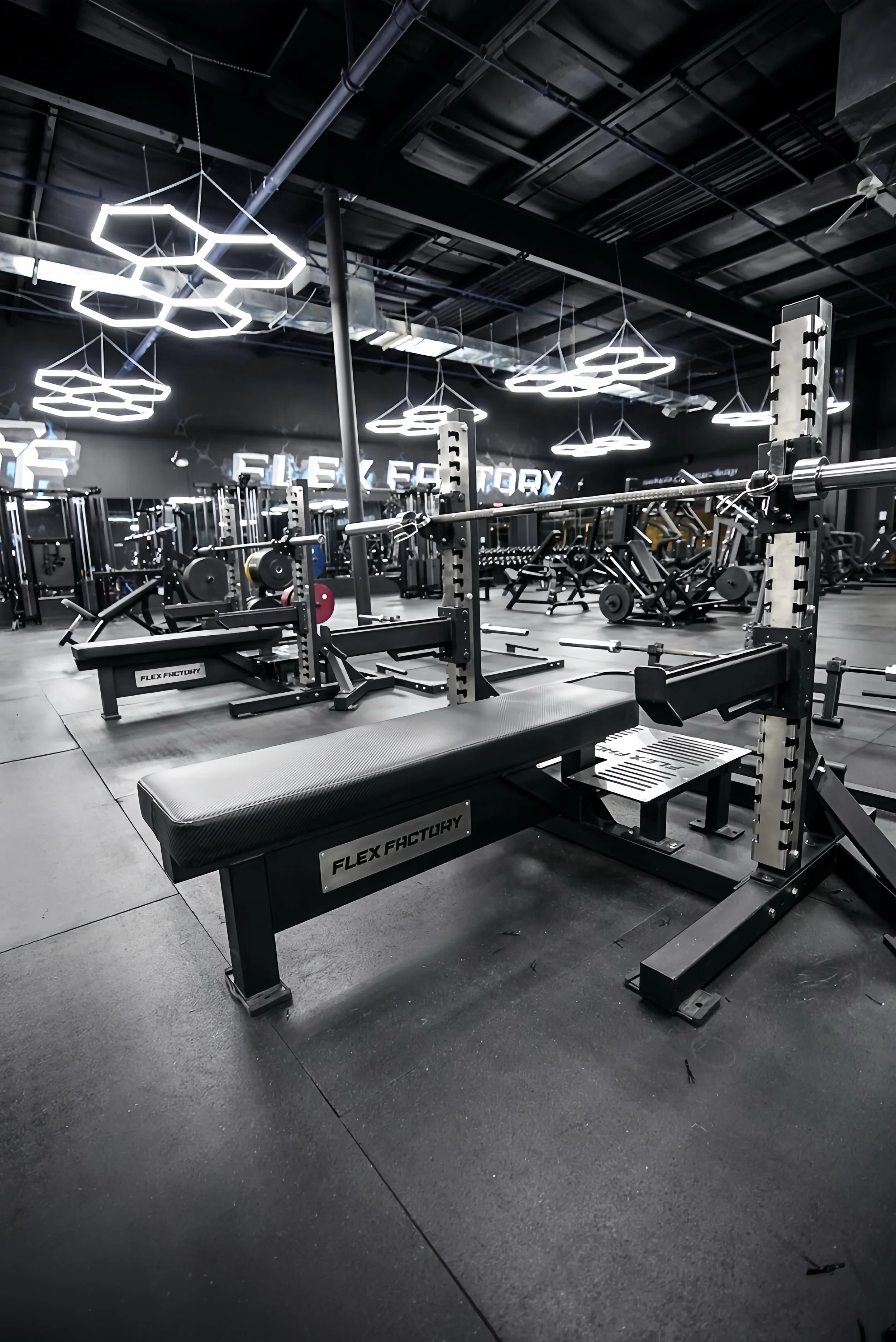 Empty gym with weightlifting equipment, benches, and racks under modern hexagon-shaped ceiling lights.