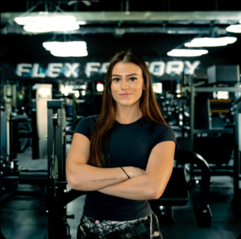 A woman standing with arms crossed in a gym with fitness equipment and a sign reading 'FLEX F' in the background.