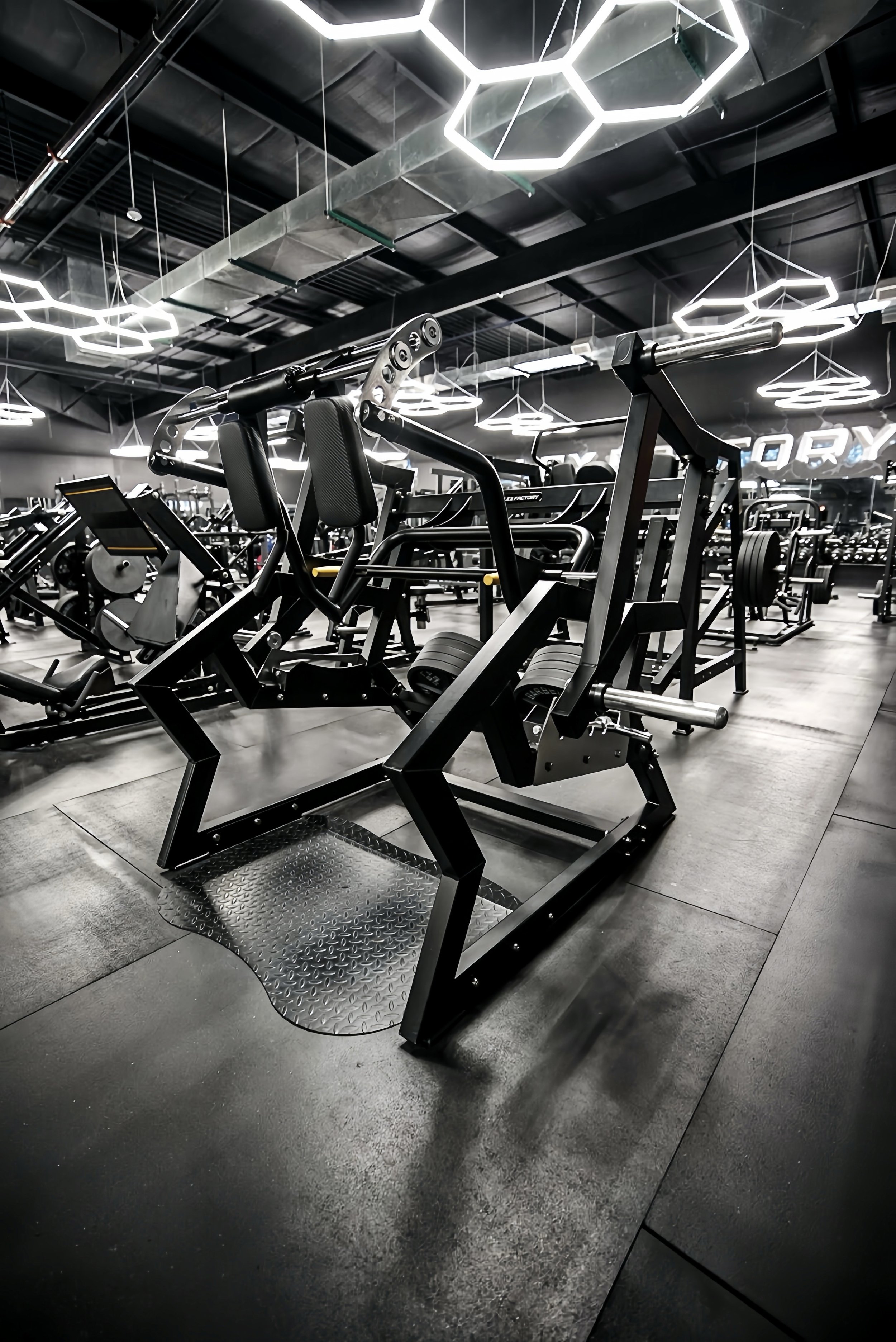 Interior of a modern gym with various exercise equipment, including a leg press machine in the foreground and illuminated hexagon light fixtures on the ceiling.