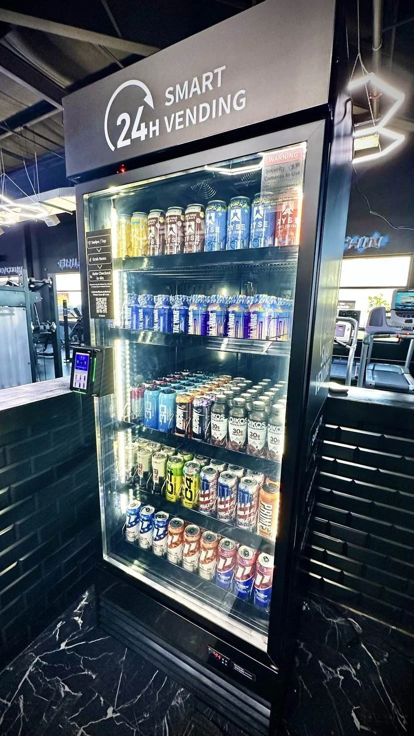 A vending machine labeled '24H SMART VENDING' stocked with energy drinks and canned beverages inside a gym or fitness center.