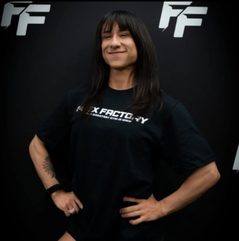 A woman with long dark hair smiling confidently, standing with her hands on her hips, wearing a black T-shirt with the text 'XFACTOR' and a dark background featuring logos.