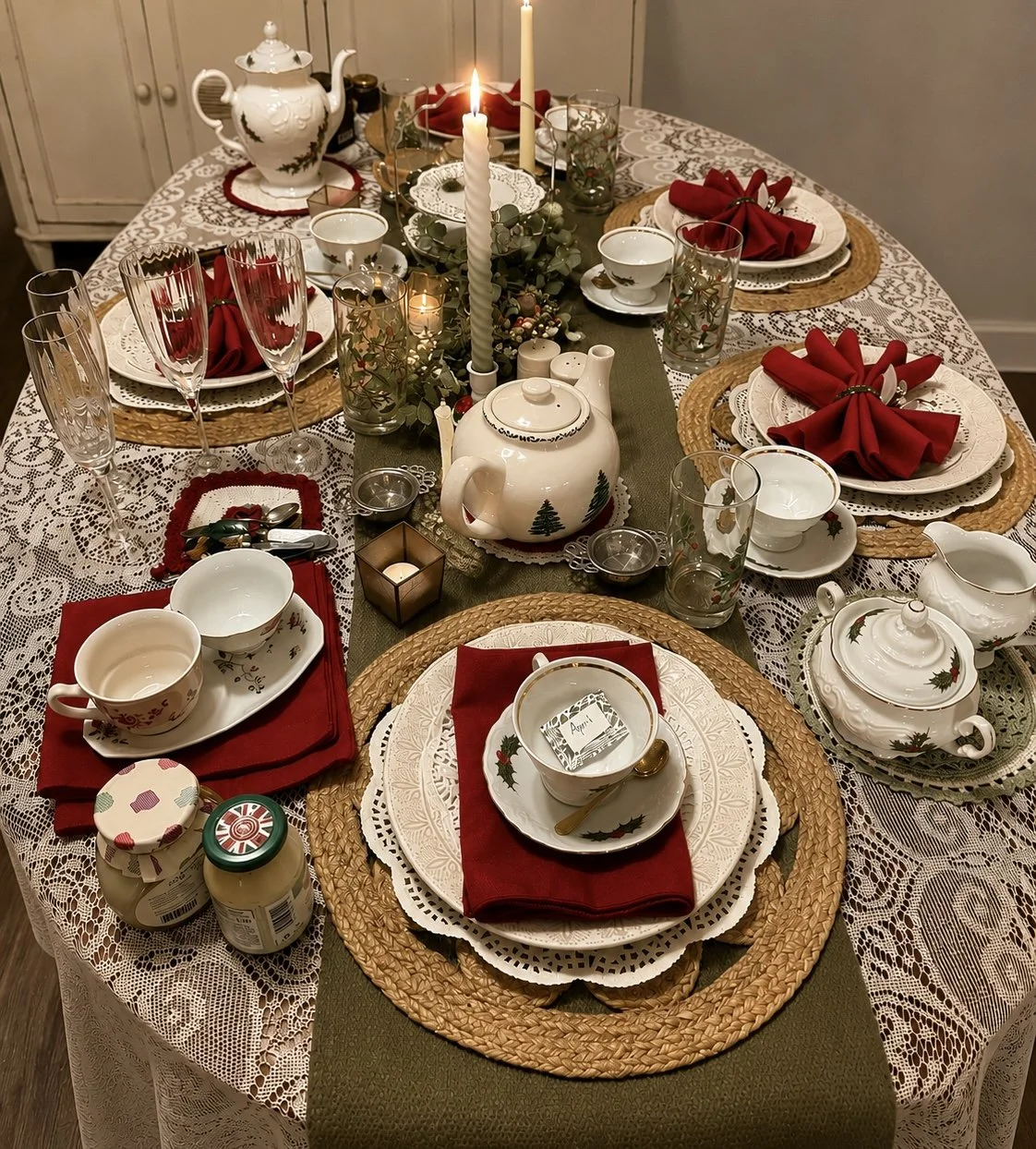 A Christmas-themed dinner table set with white and gold china, red cloth napkins, woven placemats, and a lace tablecloth. The centerpiece features candles, greenery, and a ceramic teapot decorated with holiday motifs. There are glasses, cups, and sma