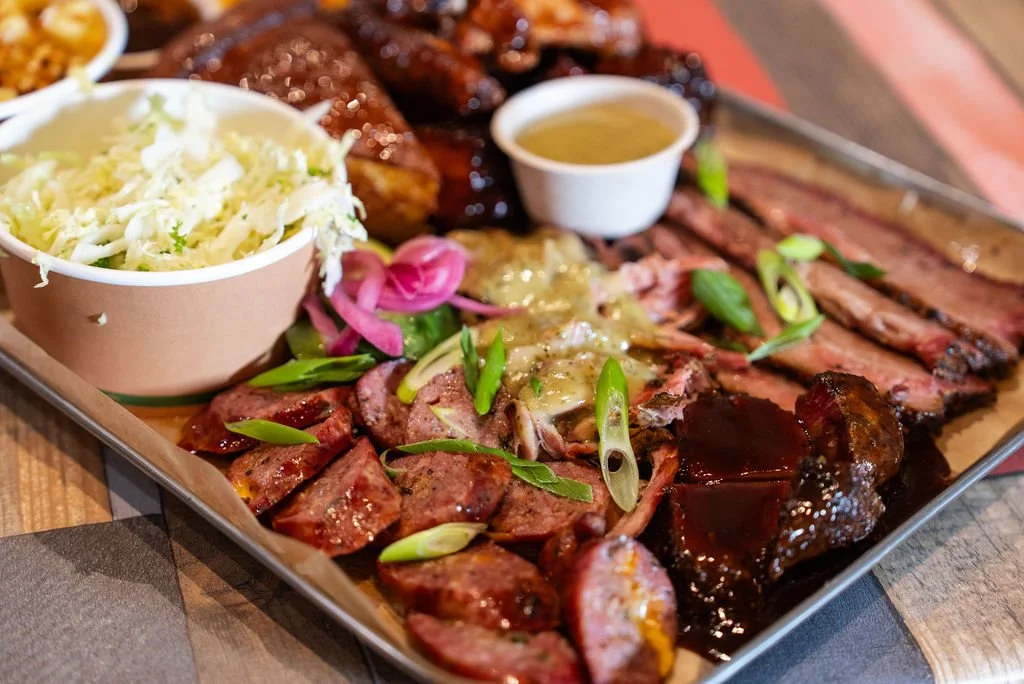 A platter of assorted barbecue meats including ribs, brisket, sausage, and chicken, garnished with green onions and served with sides of coleslaw and dipping sauces.