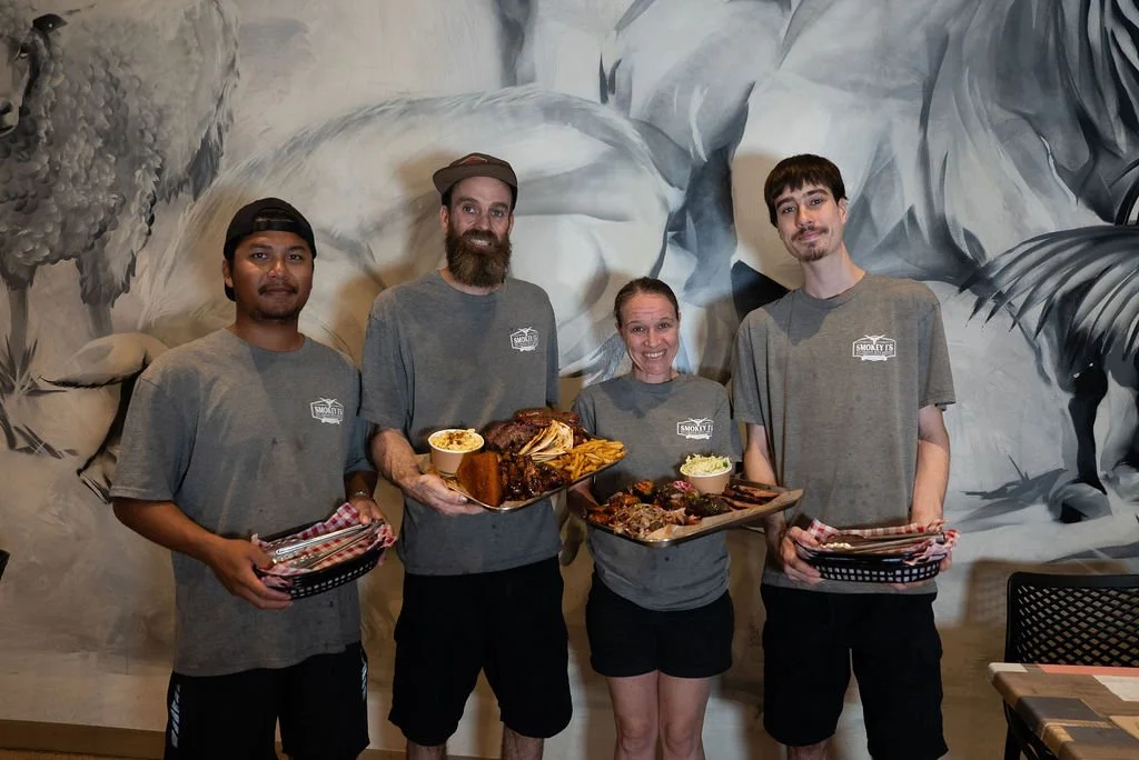 Four people standing in front of a mural with animals, holding trays of food, all wearing gray shirts. The second person from the left is holding a plate with ribs and fries, while the third person is holding a tray of meat and sides.
