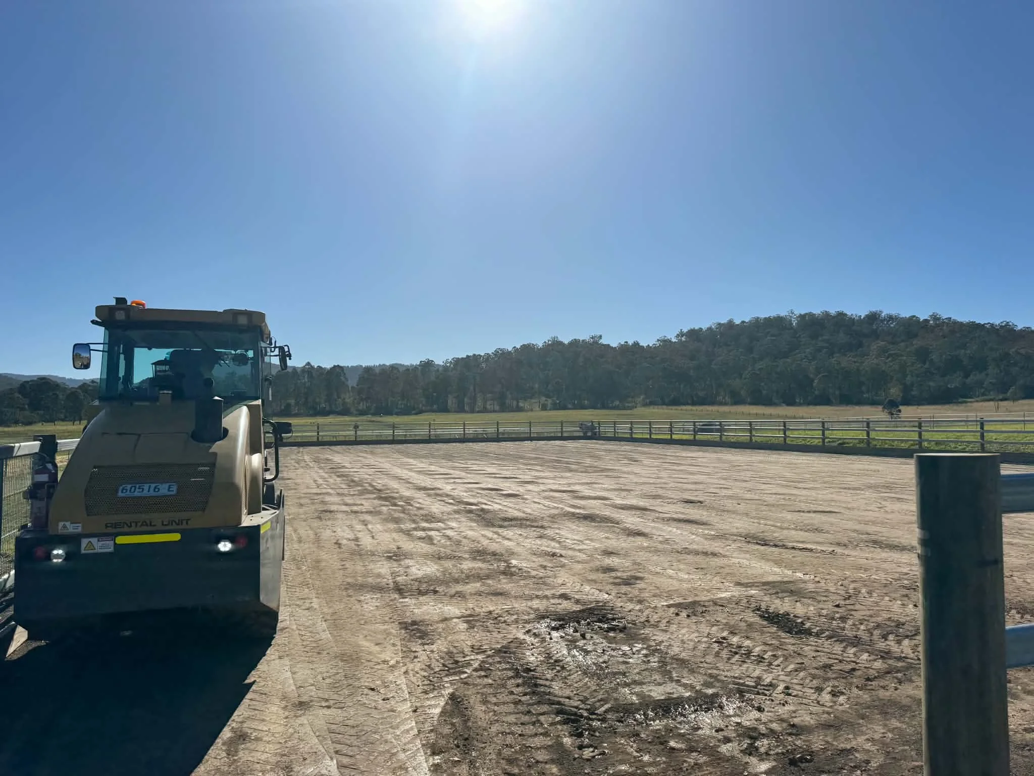 View of a blue sky with the sun shining brightly, farmland with trees and hills in the background, and a tractor in the foreground on a dirt surface.
