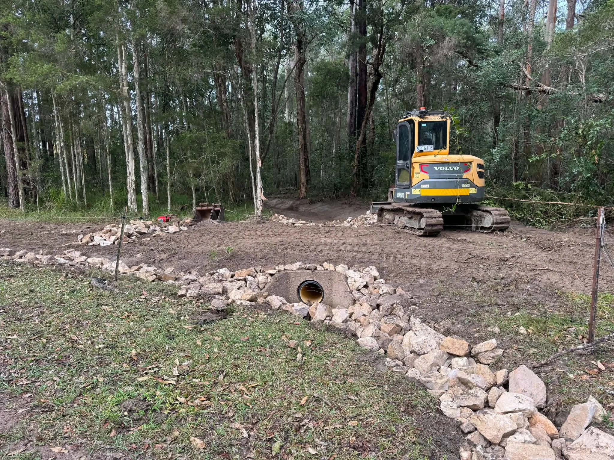 A small yellow and black excavator parked on dirt in a wooded area, with rocks placed in a line on the ground.