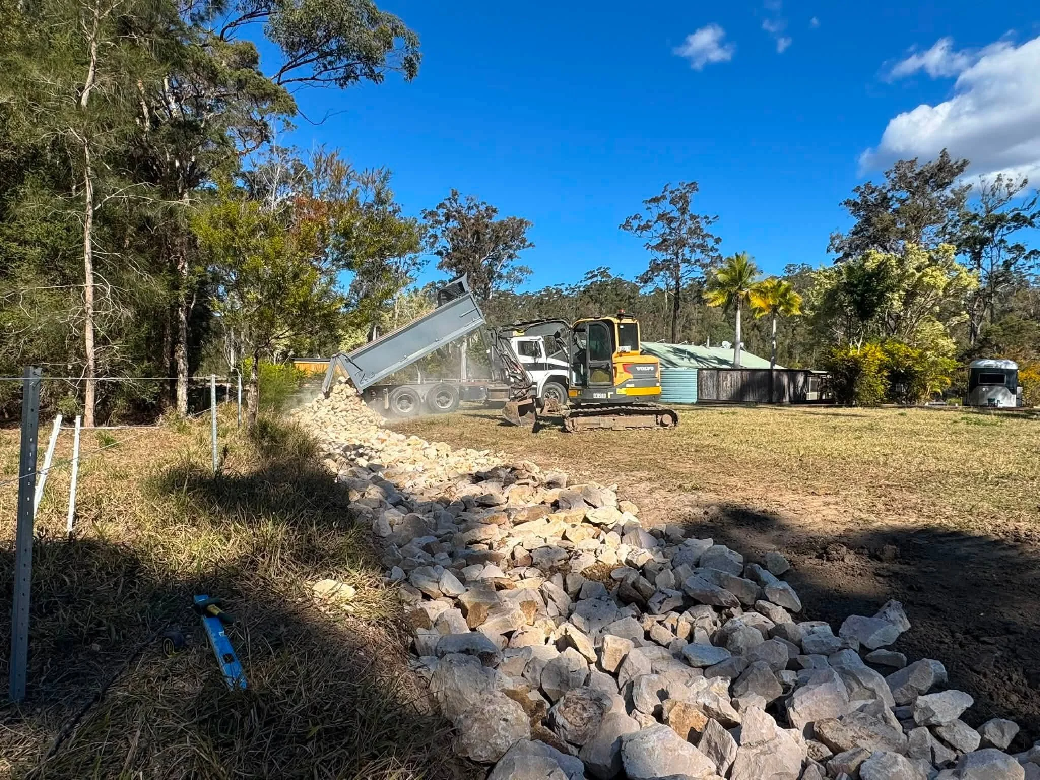 Heavy construction equipment dumping rocks onto a pile in a yard with trees and a clear blue sky.
