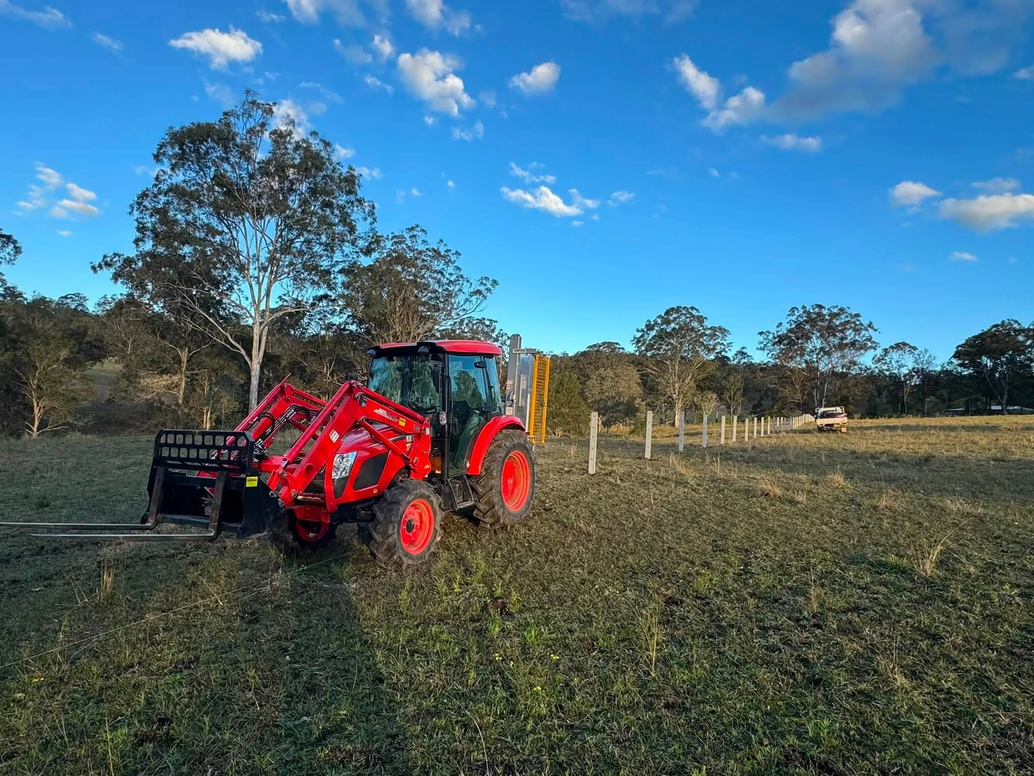 A red tractor with a front loader attachment parked on a grassy field under a blue sky with scattered clouds. There are trees and a white truck in the background.
