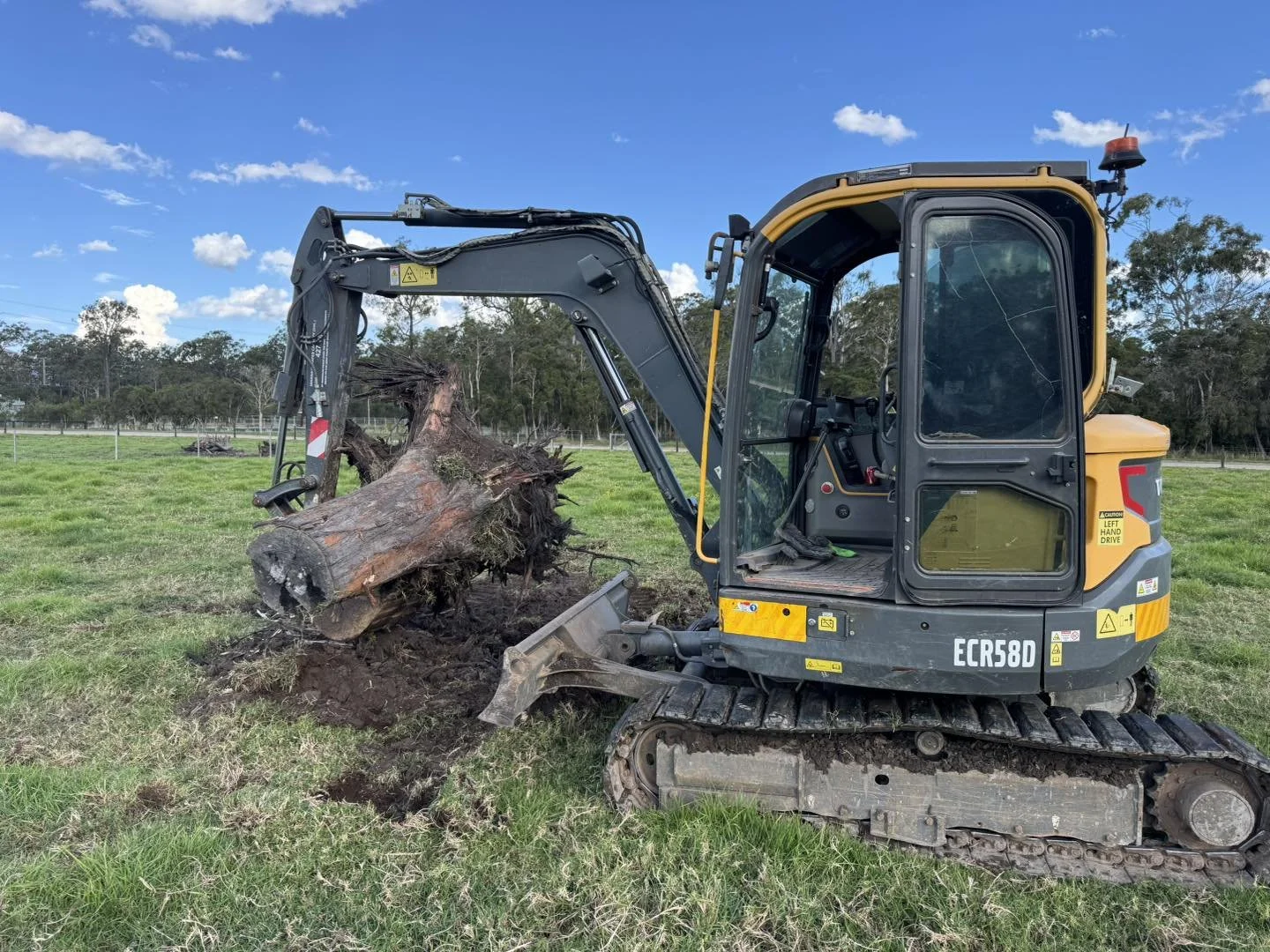 A small Caterpillar excavator uprooting a large tree stump on a grassy field with a blue sky and some clouds.