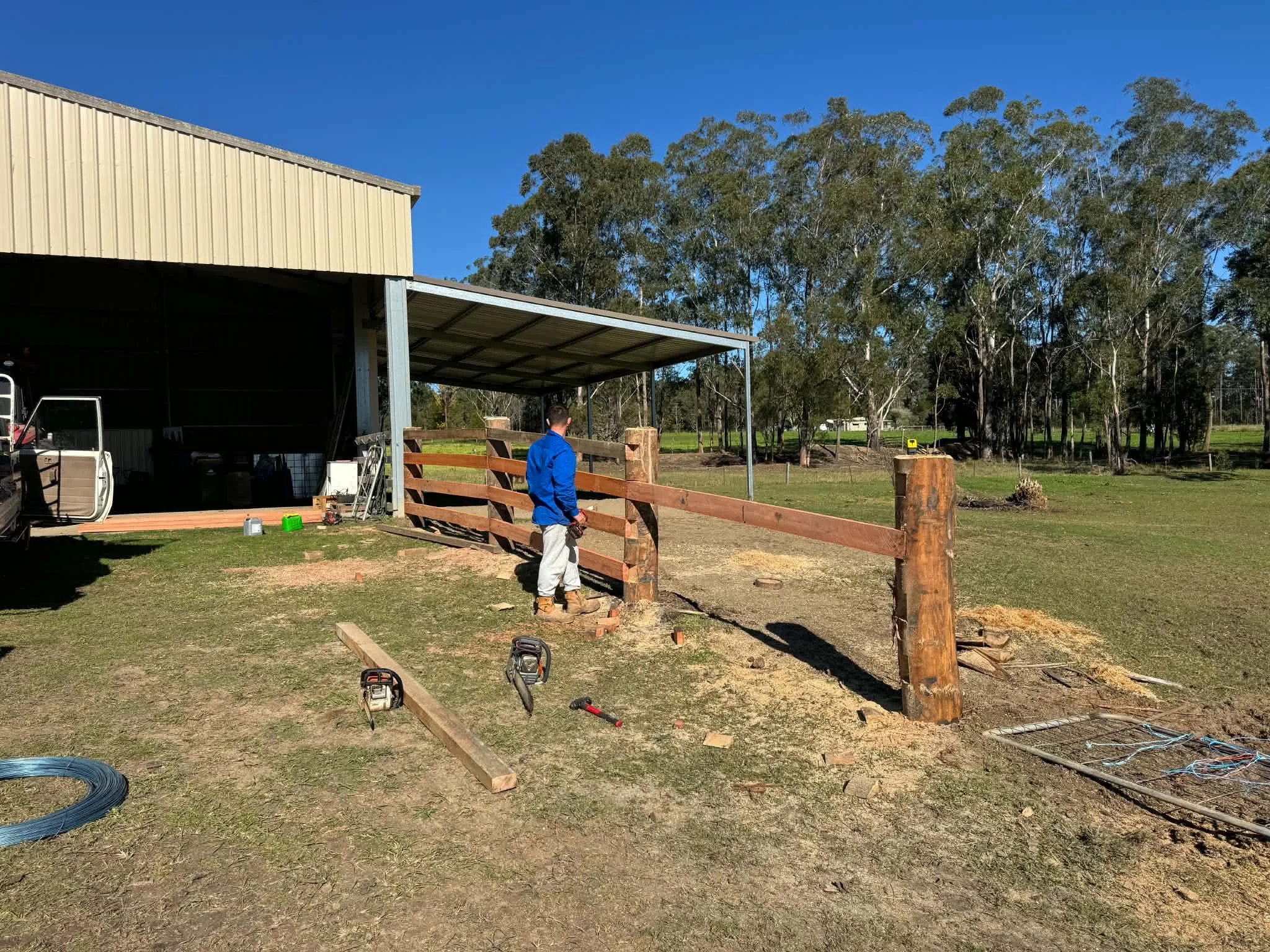 A man in a blue shirt and white pants standing outdoors near a partially built wooden fence. There is construction equipment and tools on the ground, with trees and a blue sky in the background.