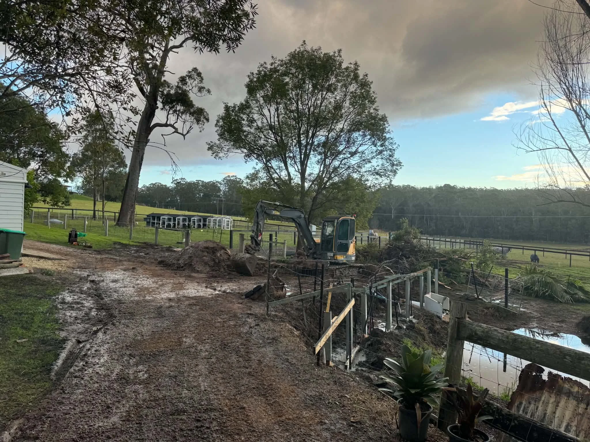 A backyard area with trees, a muddy dirt path, a small excavator digging near a water feature, and a fenced-in pond, under a cloudy sky with rain clouds and some blue sky.
