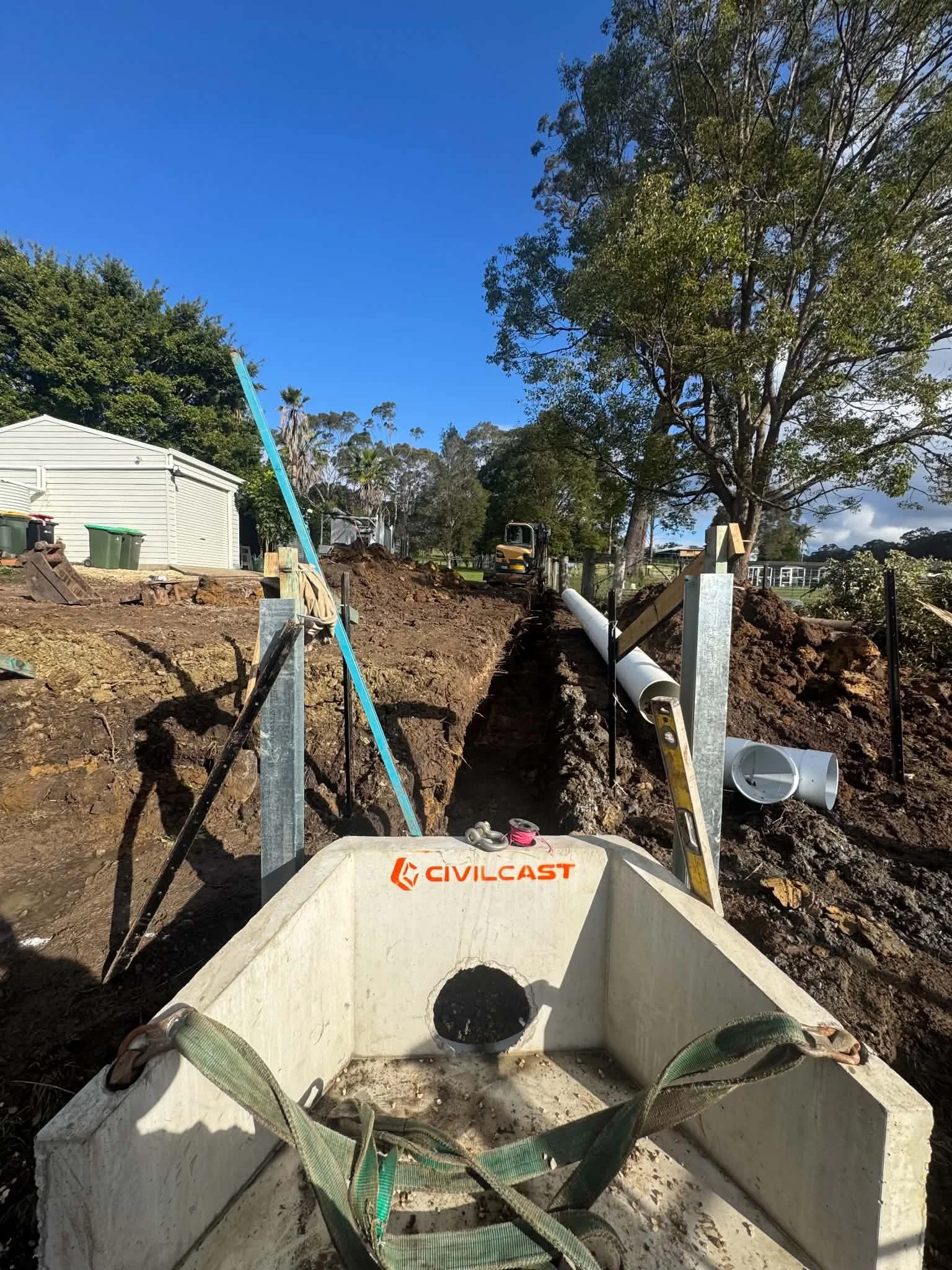 View from the front of a construction site with a large white pipe in the ground, surrounded by tools, straps, and debris. In the background, there are trees, a blue sky, and construction equipment.