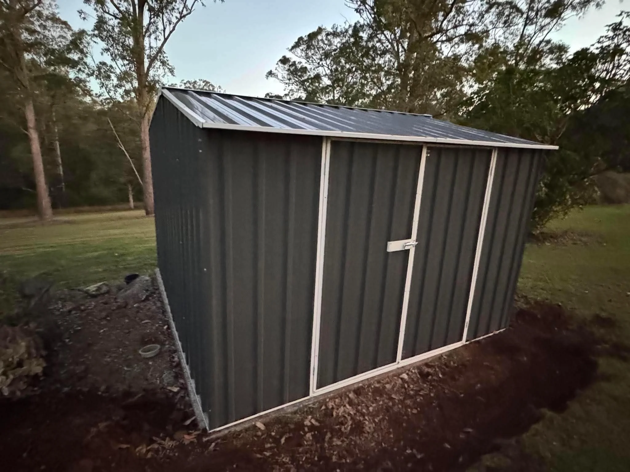 A dark gray shed with a metal roof, surrounded by trees and dirt ground in a natural outdoor setting.
