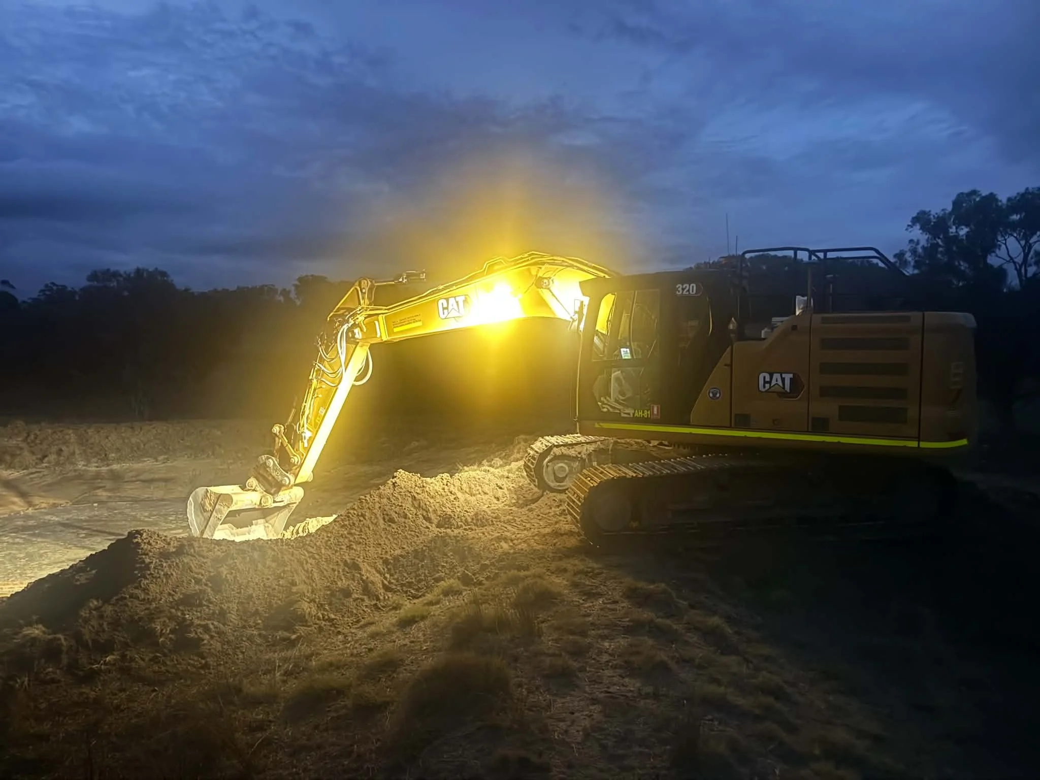 Construction excavator digging soil at dusk, illuminated by bright yellow lights, with a dark cloudy sky and trees in the background.