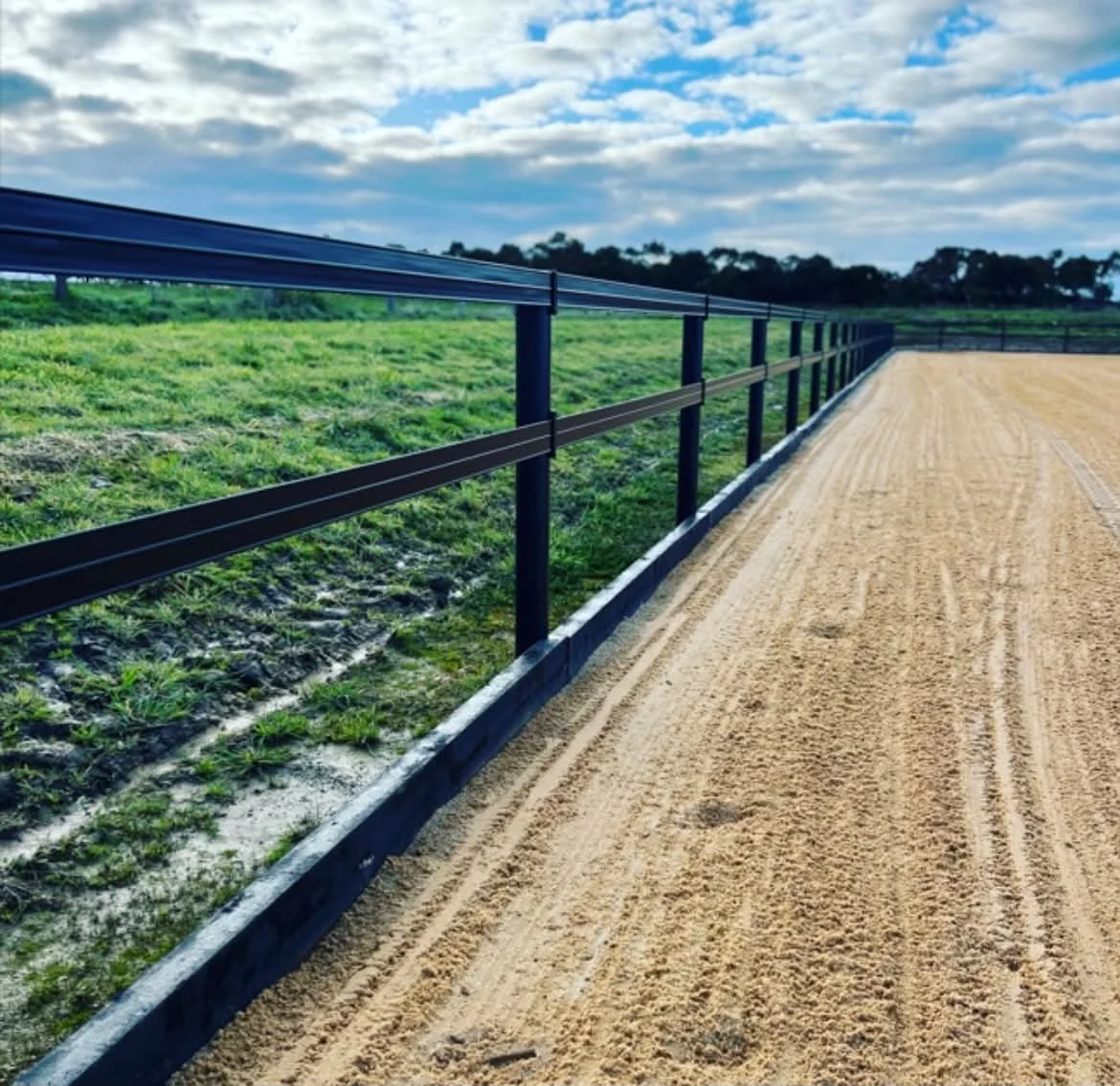 View of a dirt racetrack with a black metal fence on the left side and a grassy field beyond, under a partly cloudy sky.