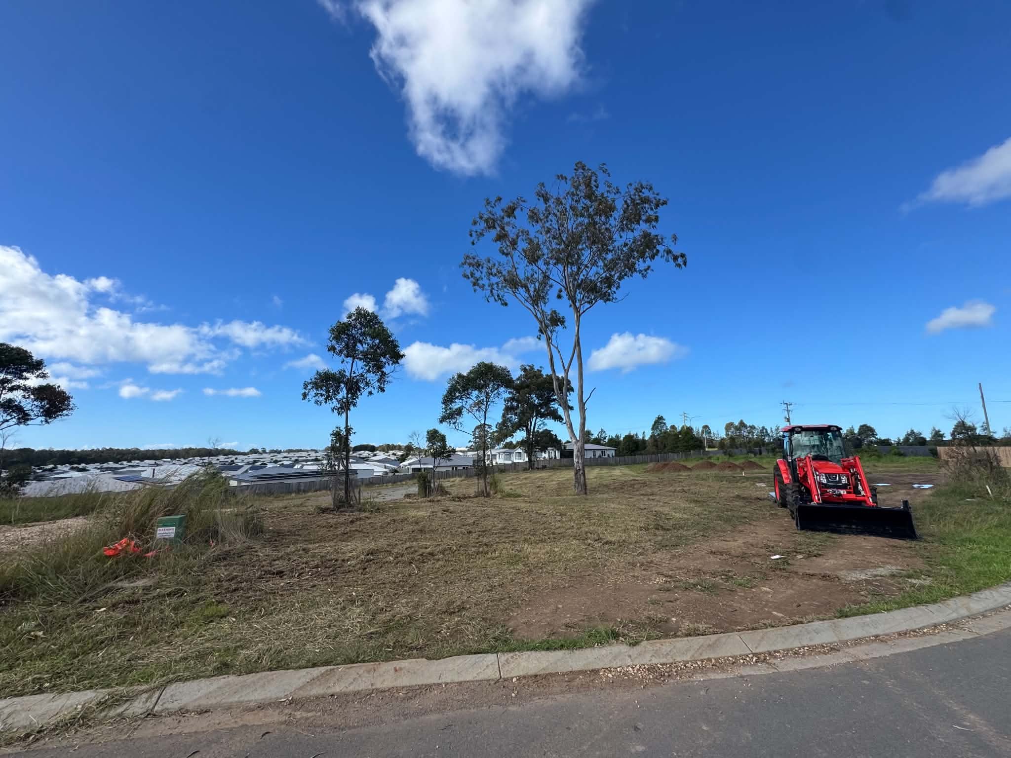 Construction land with the front view of a red tractor, trees, a row of houses, clear sky with some clouds, and power lines in the distance.