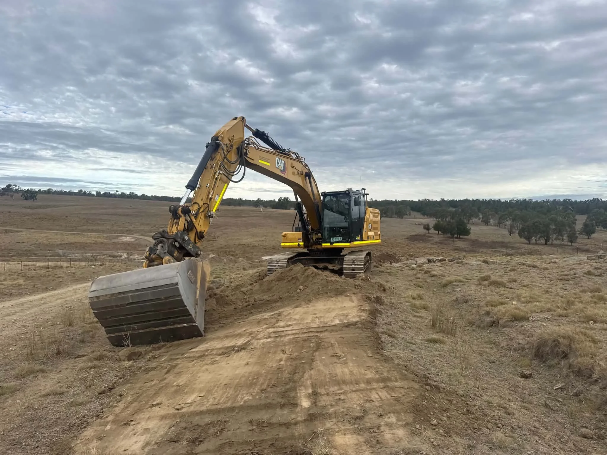 A large yellow excavator with a Caterpillar logo is moving dirt on an open construction site, with a cloudy sky overhead and distant trees on the horizon.