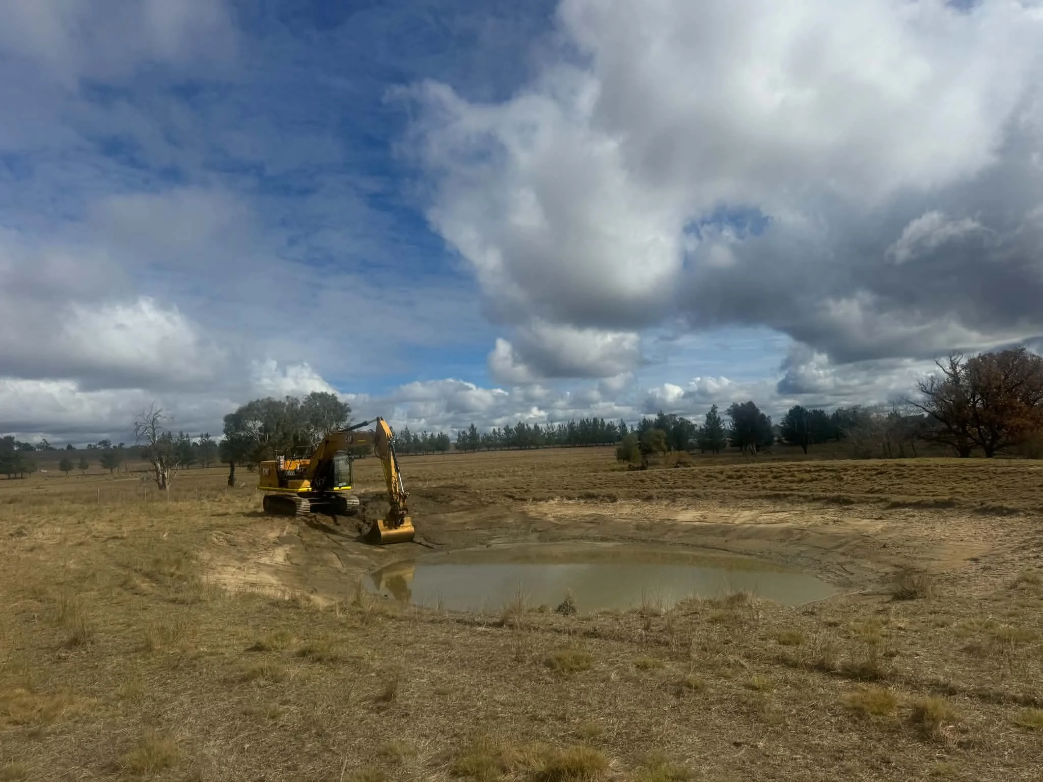 A rural landscape scene with a yellow excavator working on a field, surrounded by trees under a partly cloudy sky.