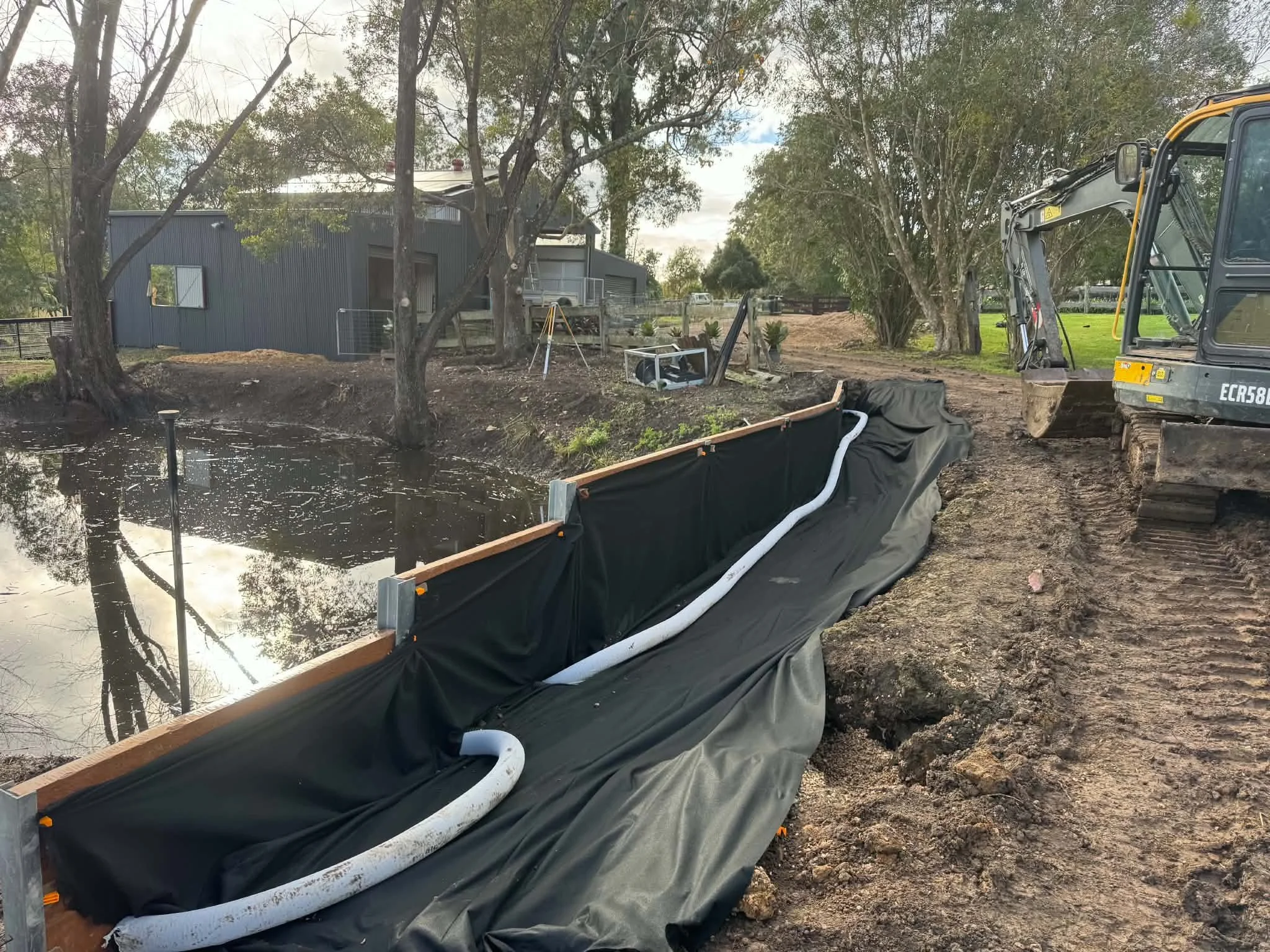Construction site with a pond, trees, and a building in the background. A large excavator is parked on the right side, and there is a black liner or fabric along the edge of the pond, with white pipes visible. The ground is muddy.
