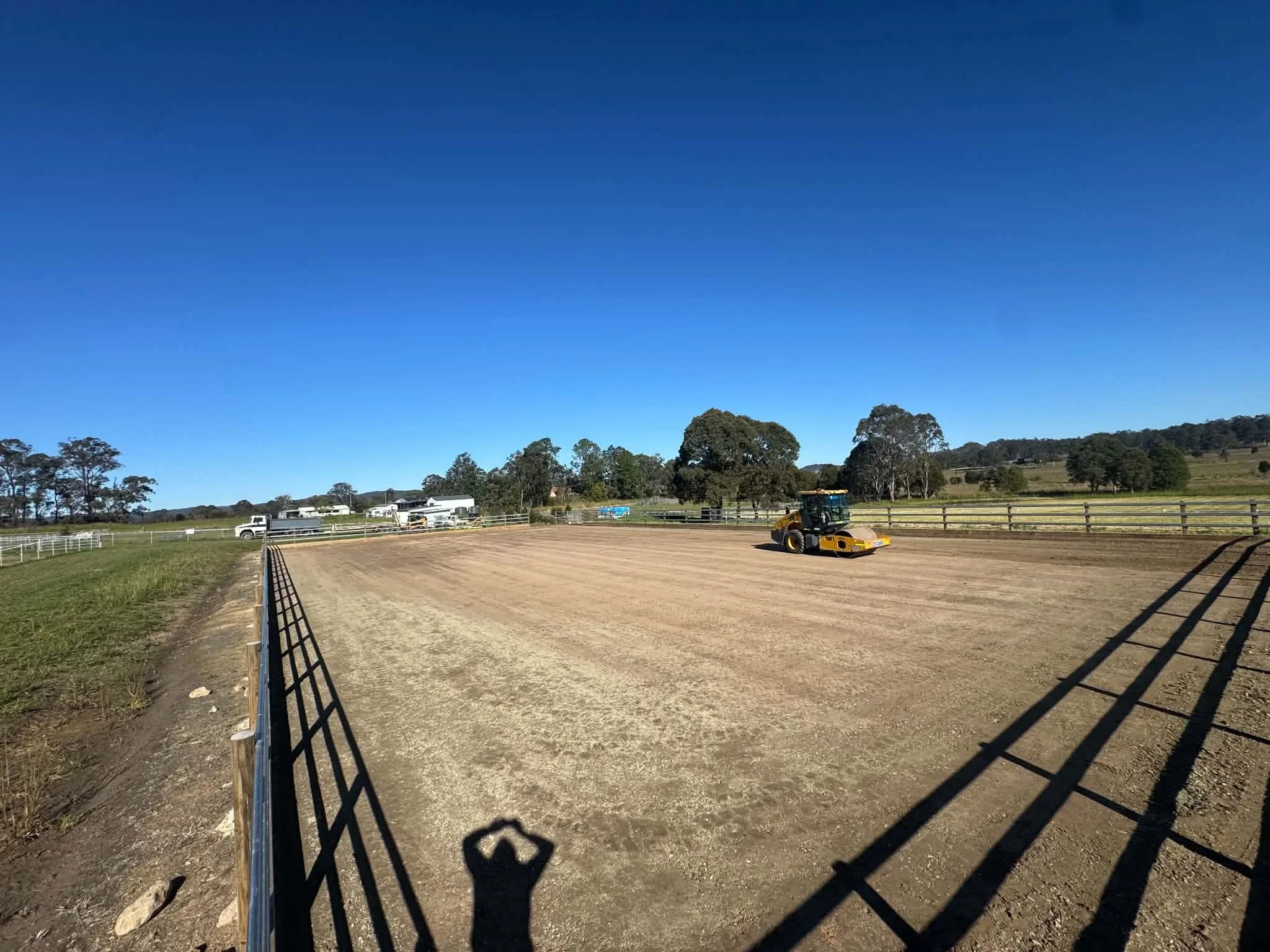A dirt race track with a roller on it, surrounded by a black railing and fencing, empty in the foreground. In the background, there are trees, buildings, and a tractor under a clear blue sky.