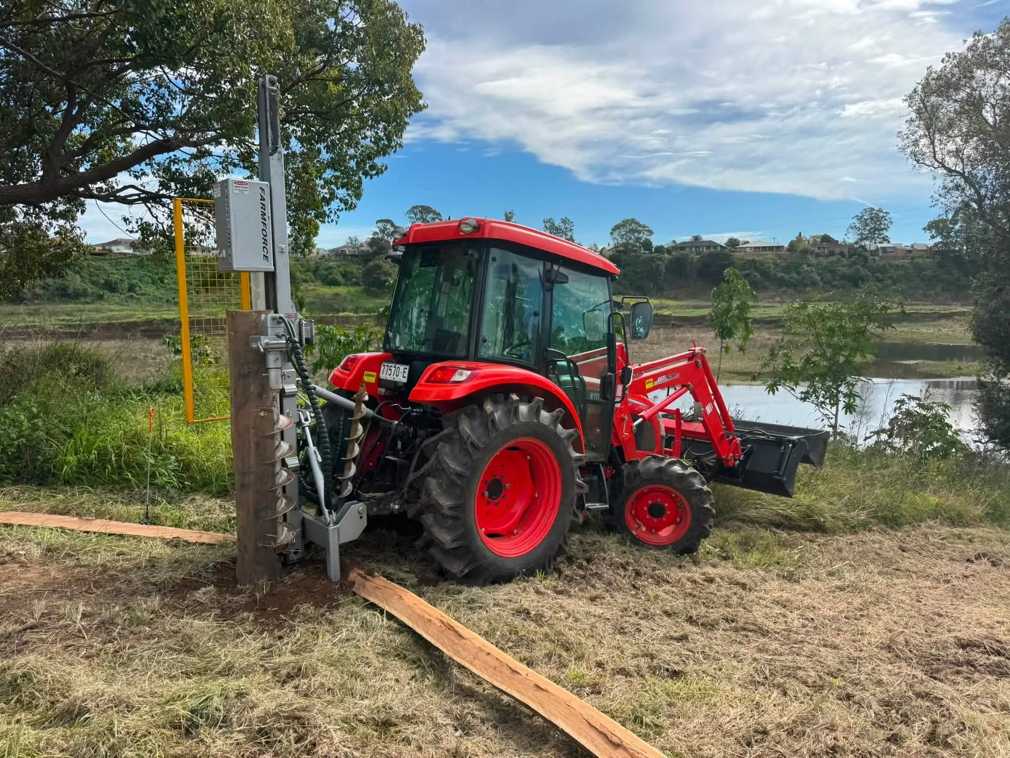 Red tractor operating near a body of water with green trees and houses in the background, under a partly cloudy sky.