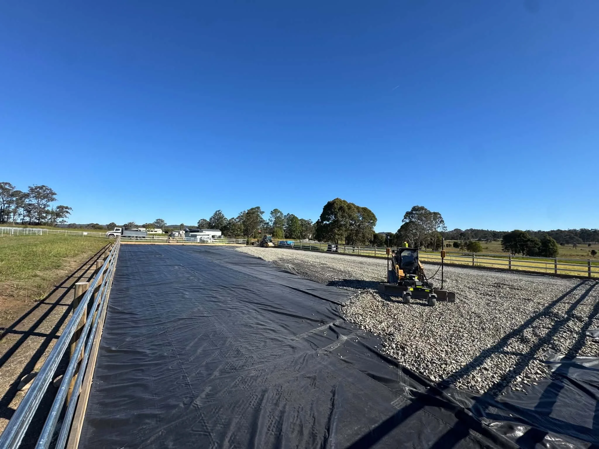 Farm with a wide blue sky, trees in the background, and a section of land under construction with black landscaping fabric and construction equipment.