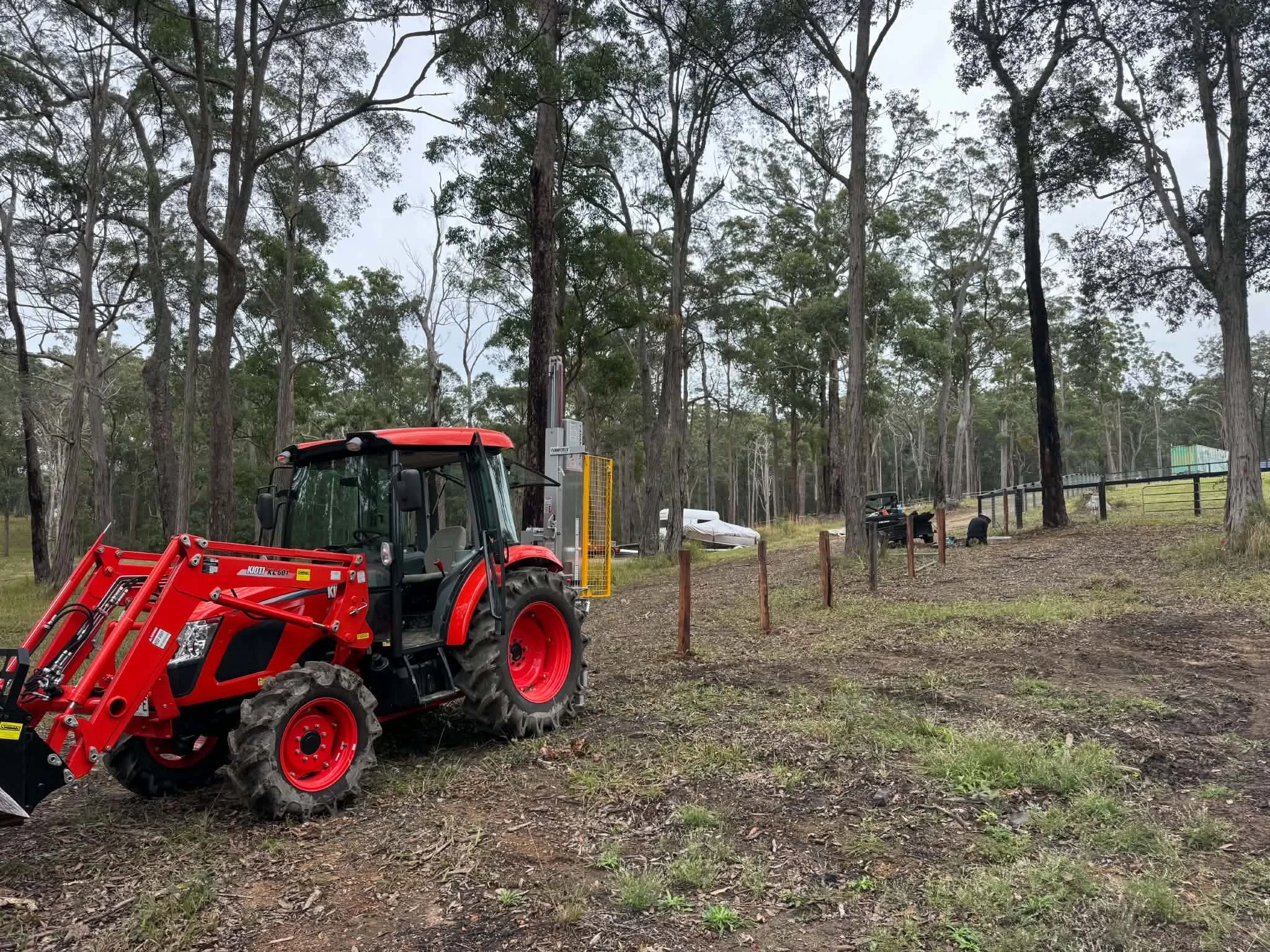 A red tractor with front loader attachments parked on a dirt ground in a wooded area with tall trees and a cloudy sky.
