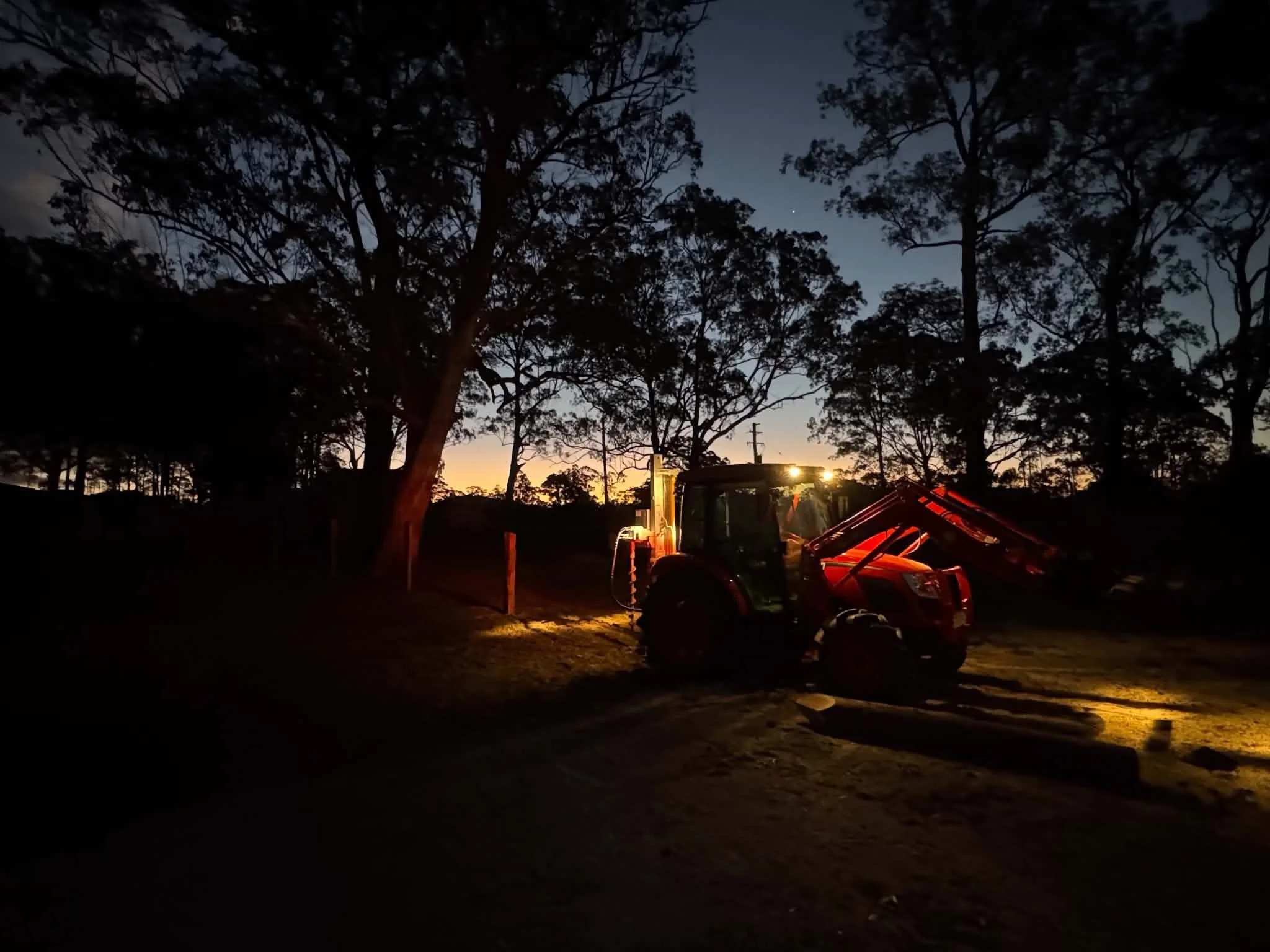 A tractor parked outdoors at dusk or dawn with trees and power lines in the background, illuminated by artificial light.