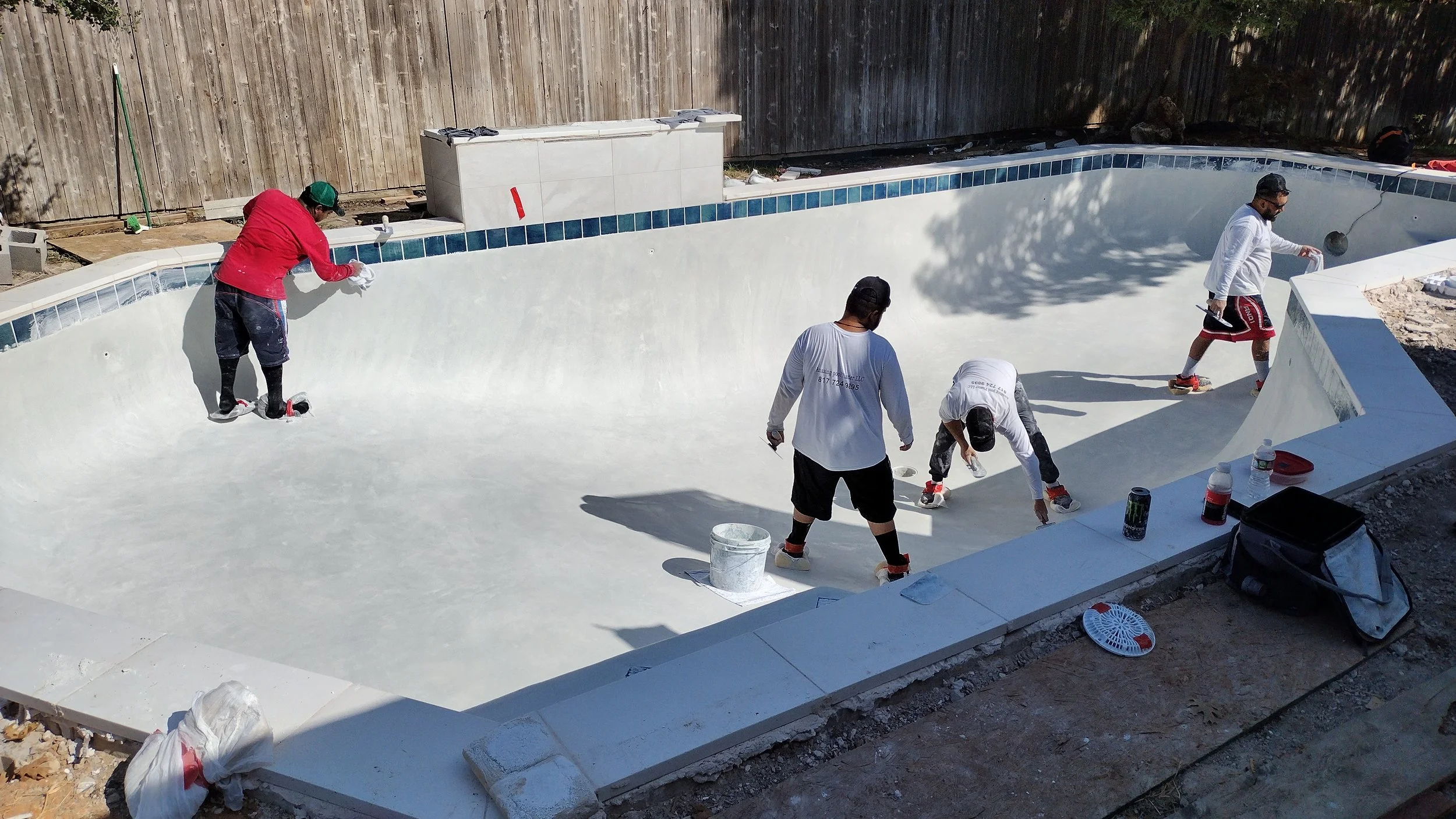 Workers renovating an empty swimming pool with white interior and blue tiles at the top edge, using tools and paint in a backyard.
