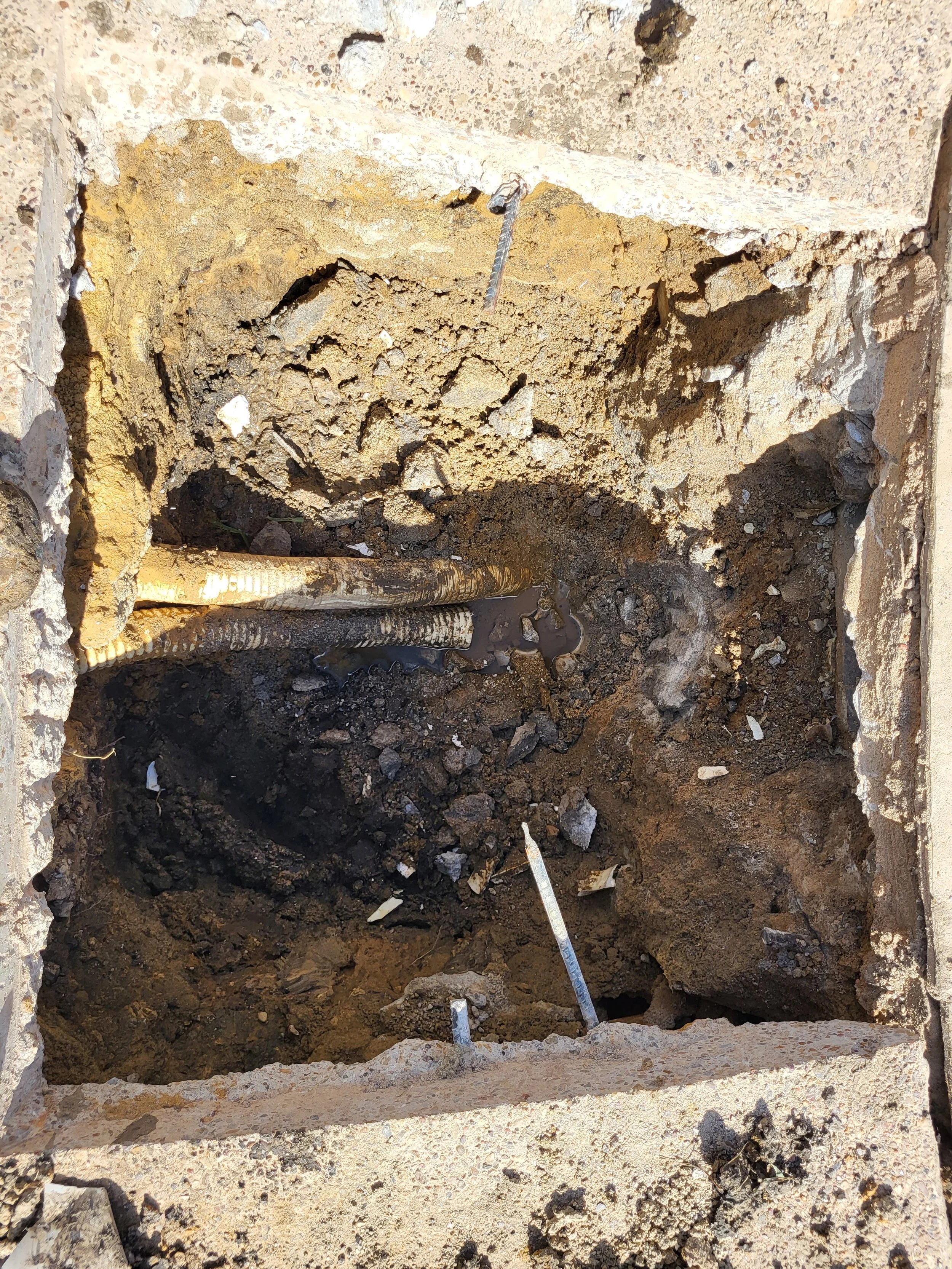 Construction site trench with exposed soil, pipes, and a measuring rod.