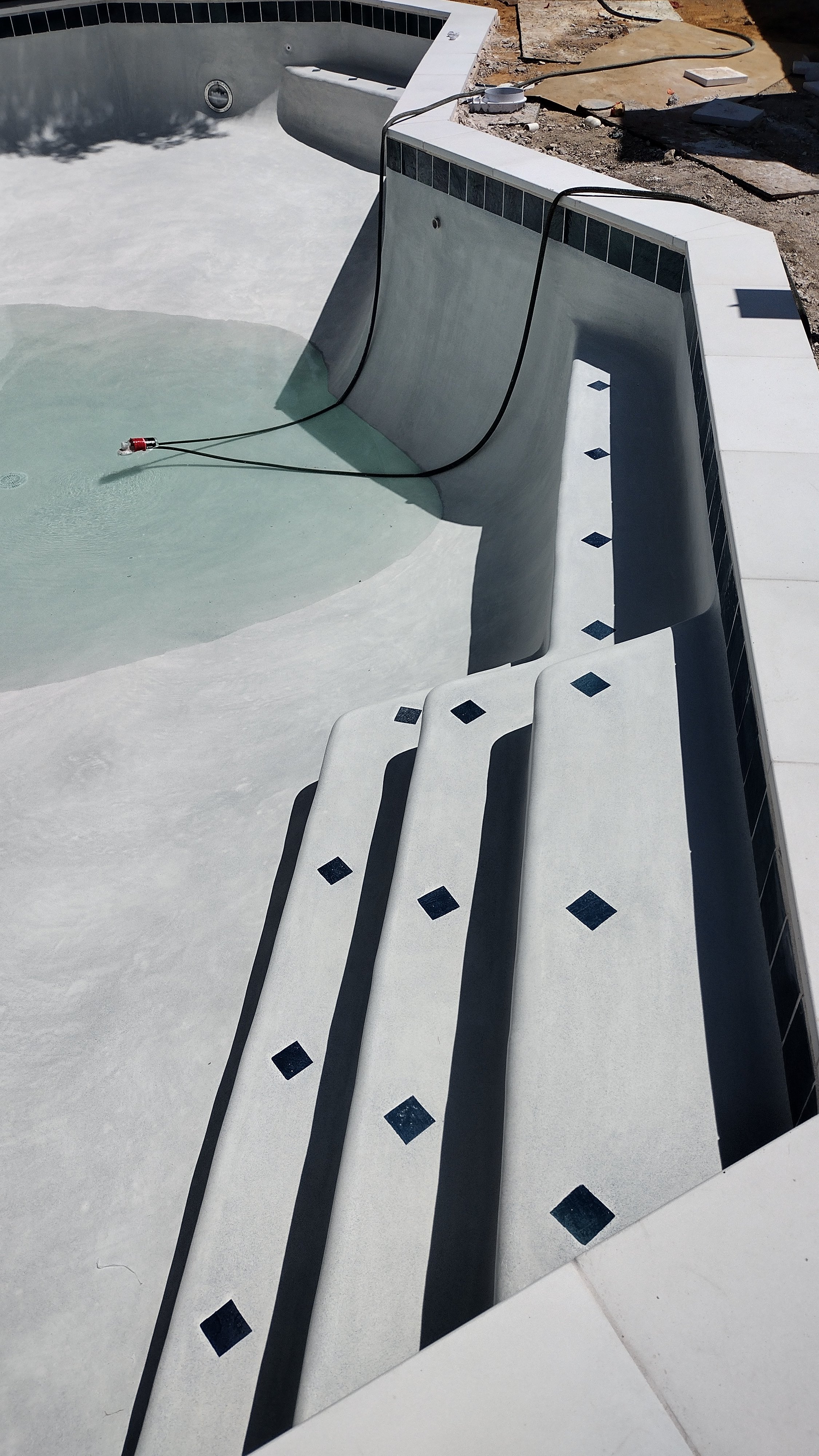 Empty swimming pool with a hydraulic lift and steps, construction materials, and a dirt surface surrounding it.