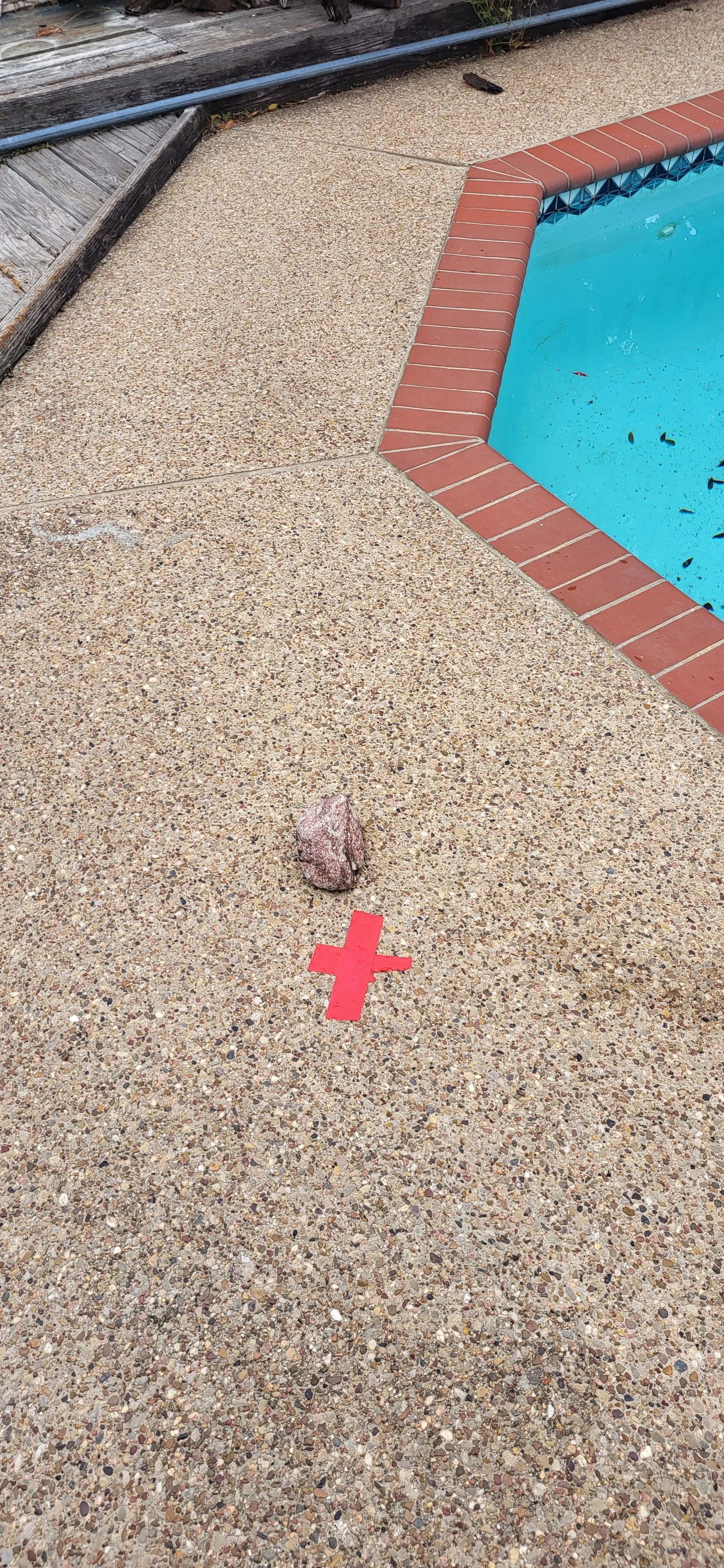 A red cross marking on a pebble-strewn concrete surface near an empty swimming pool with a red brick border.