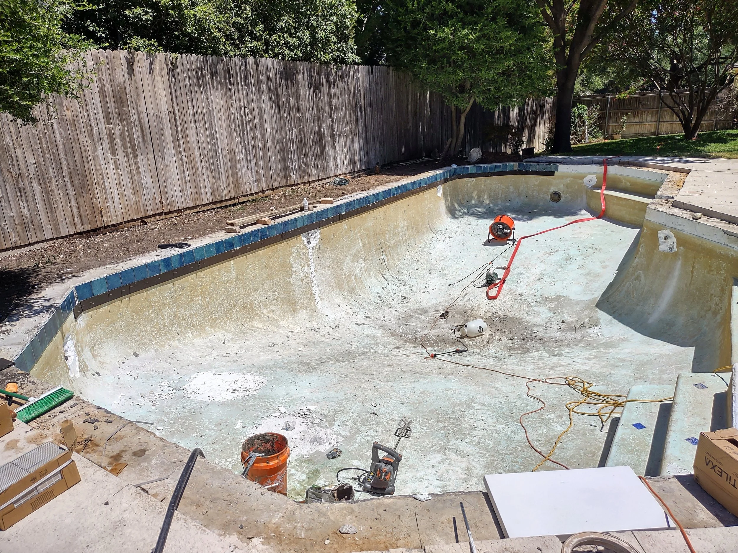 An empty swimming pool undergoing renovations with visible plumbing and construction tools around the pool area, and a wooden fence and trees in the background.