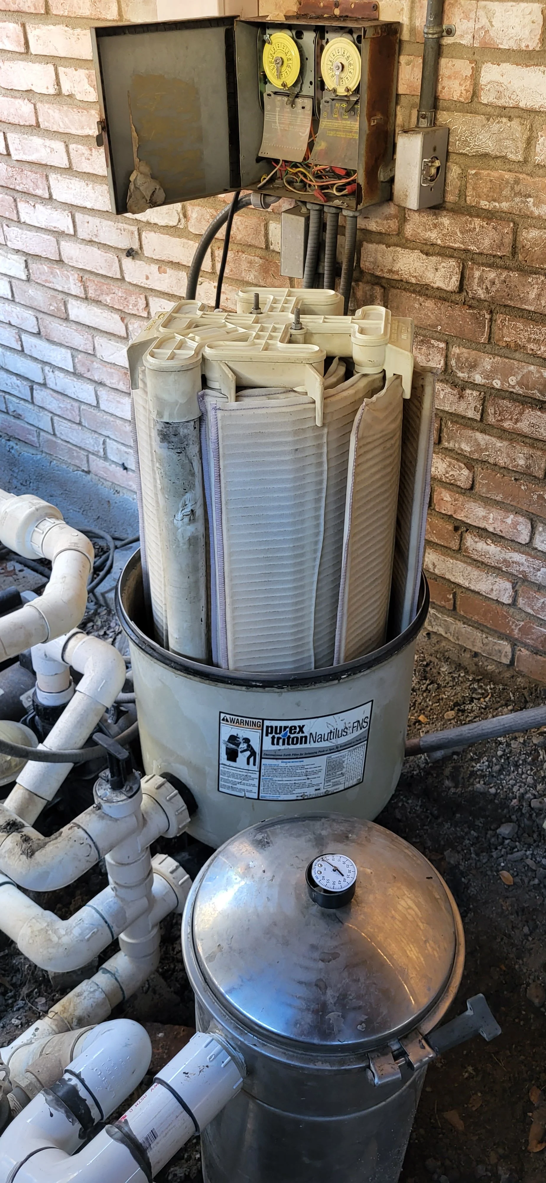 An outdoor pool filtration system with a large, cylindrical filter housing labeled "Purex Triton Nautilus FNS," connected to various white PVC pipes and valves, with a control box on a brick wall.