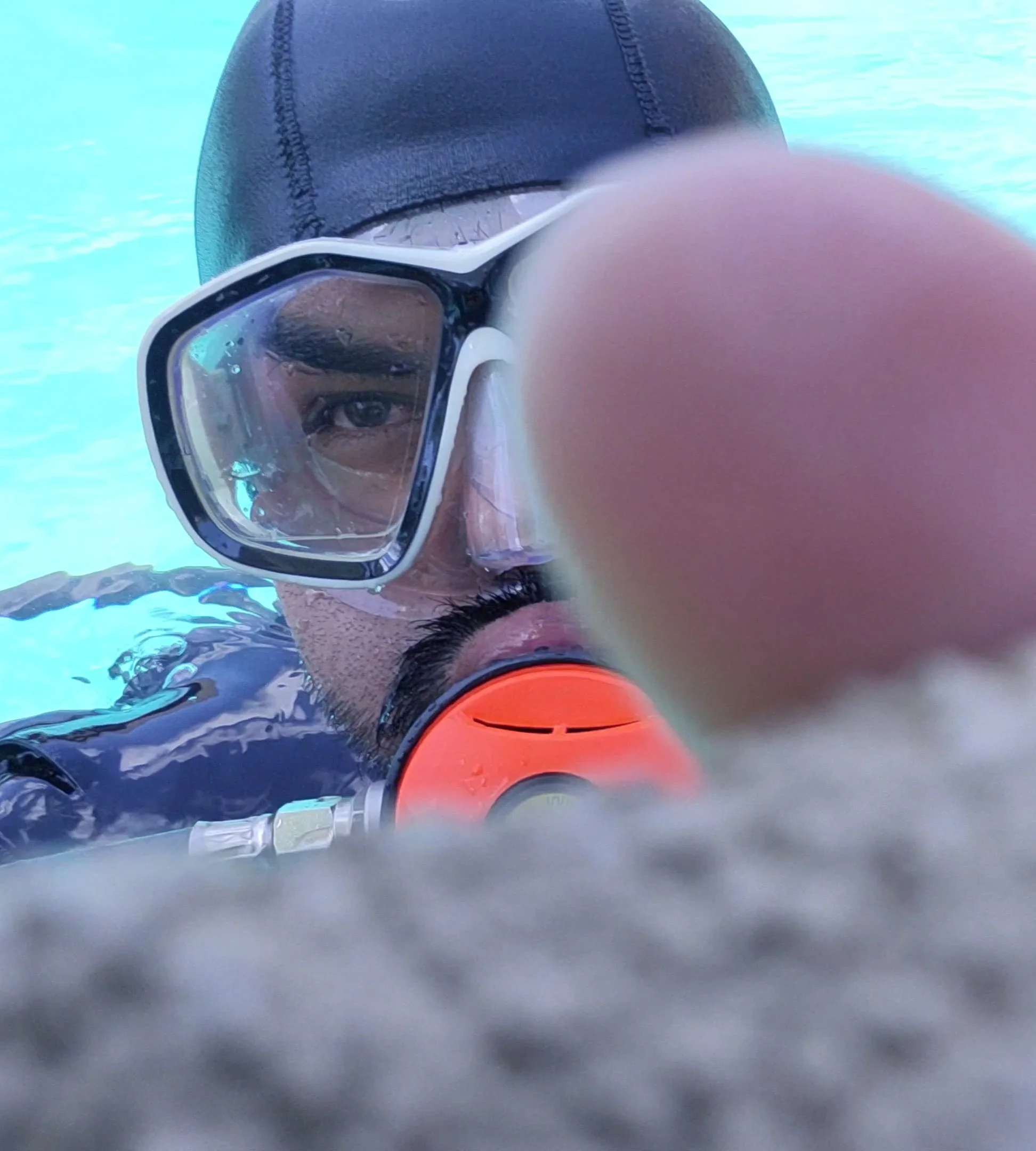 A person wearing swimming goggles and a swim cap, taking a close-up selfie underwater in a swimming pool.