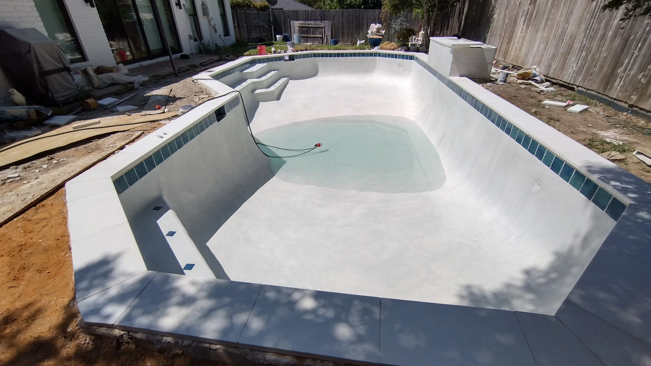 Empty swimming pool in the backyard under construction, with tools and construction materials around, and a wooden fence in the background.