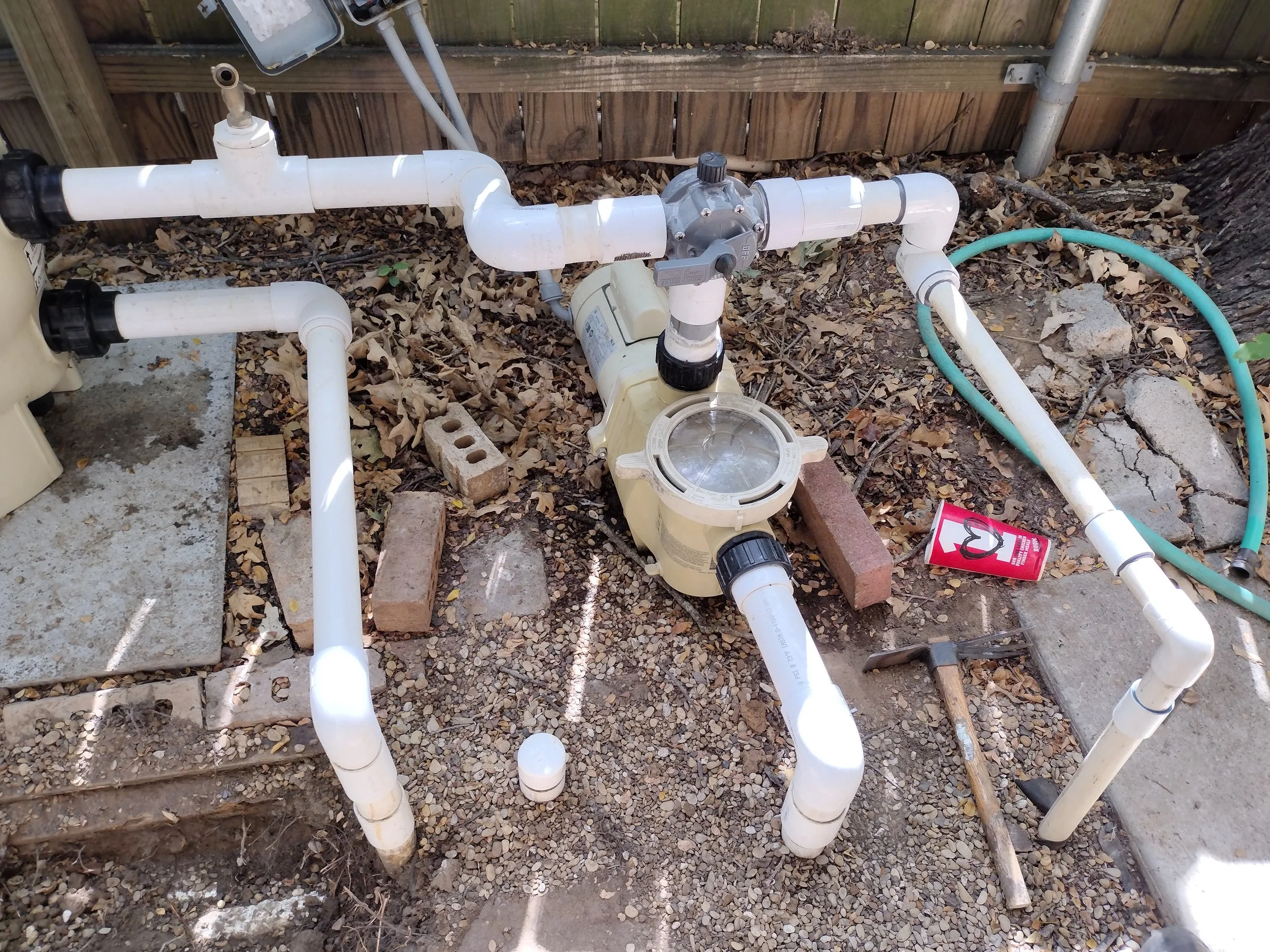 A network of white PVC pipes and a control valve installed outdoors on the ground, surrounded by gravel, leaves, and wood pieces, with a wooden fence in the background.