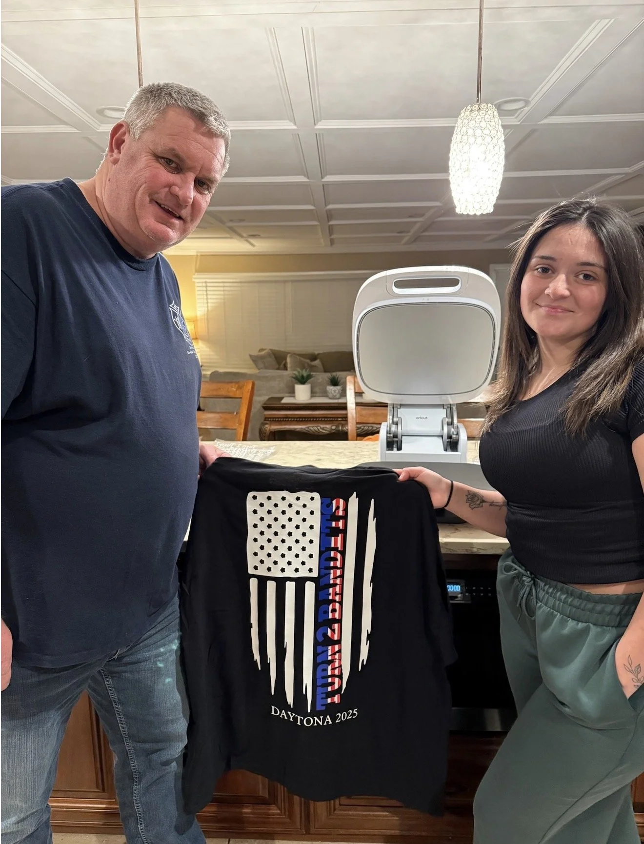Two people standing in a kitchen, holding a black themed t-shirt with a white graphic. One person is a man wearing a dark t-shirt and jeans, the other a woman in a black top and green pants. There is an open toaster oven in the background. The setting appears to be a warm-lit, cozy home.
