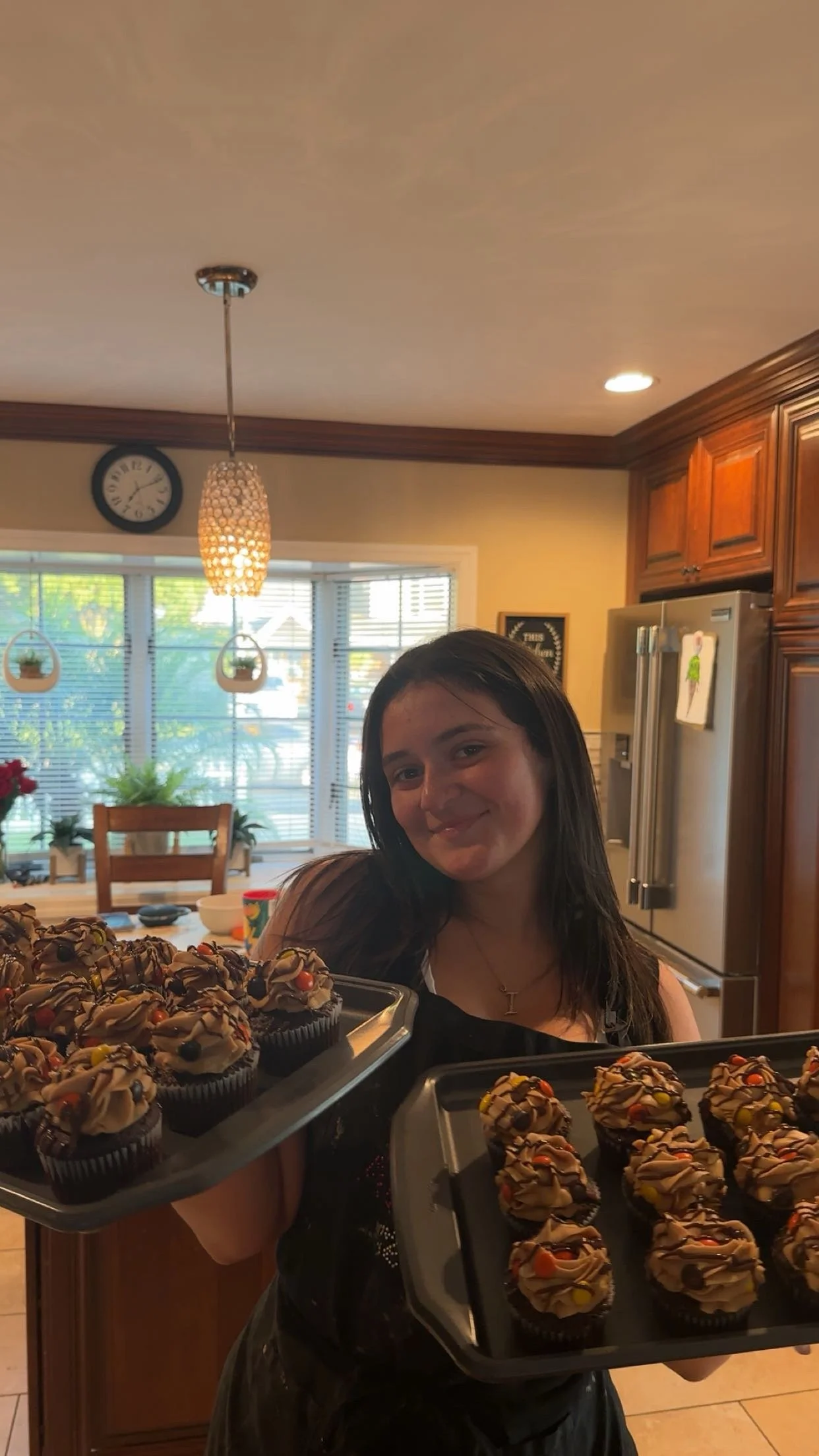 A young woman with long dark hair smiling and holding trays of chocolate cupcakes decorated with sprinkles inside a kitchen with wooden cabinets, stainless steel refrigerator, large window, and hanging light fixtures.