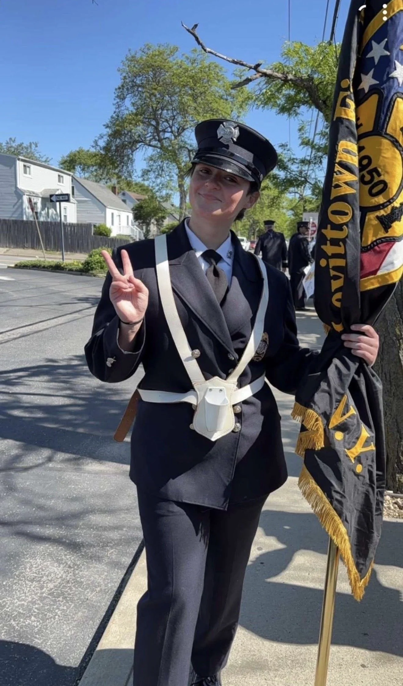 A young female police officer standing on a sidewalk, making a peace sign with her right hand, holding a flag in her left hand, and wearing a police uniform, including a hat with a badge. The background features trees, houses, and a clear blue sky.