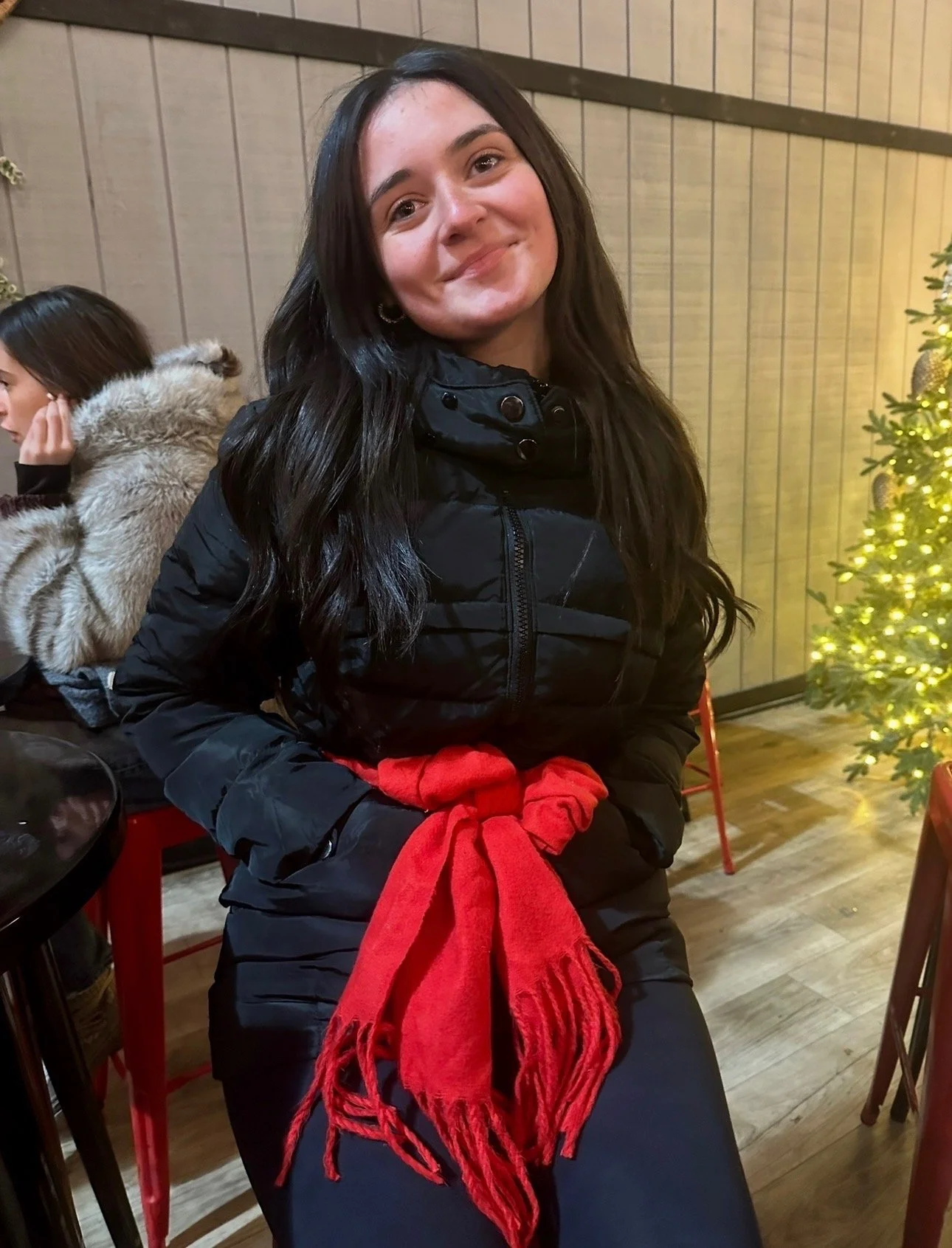 Young woman with long dark hair smiling, wearing a black jacket and a red scarf, sitting indoors near a decorated Christmas tree with lights.