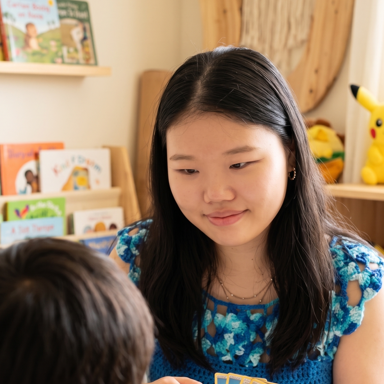 A woman with long dark hair and hoop earrings smiling while interacting with children in a room decorated with children's books and a Pikachu plush toy.