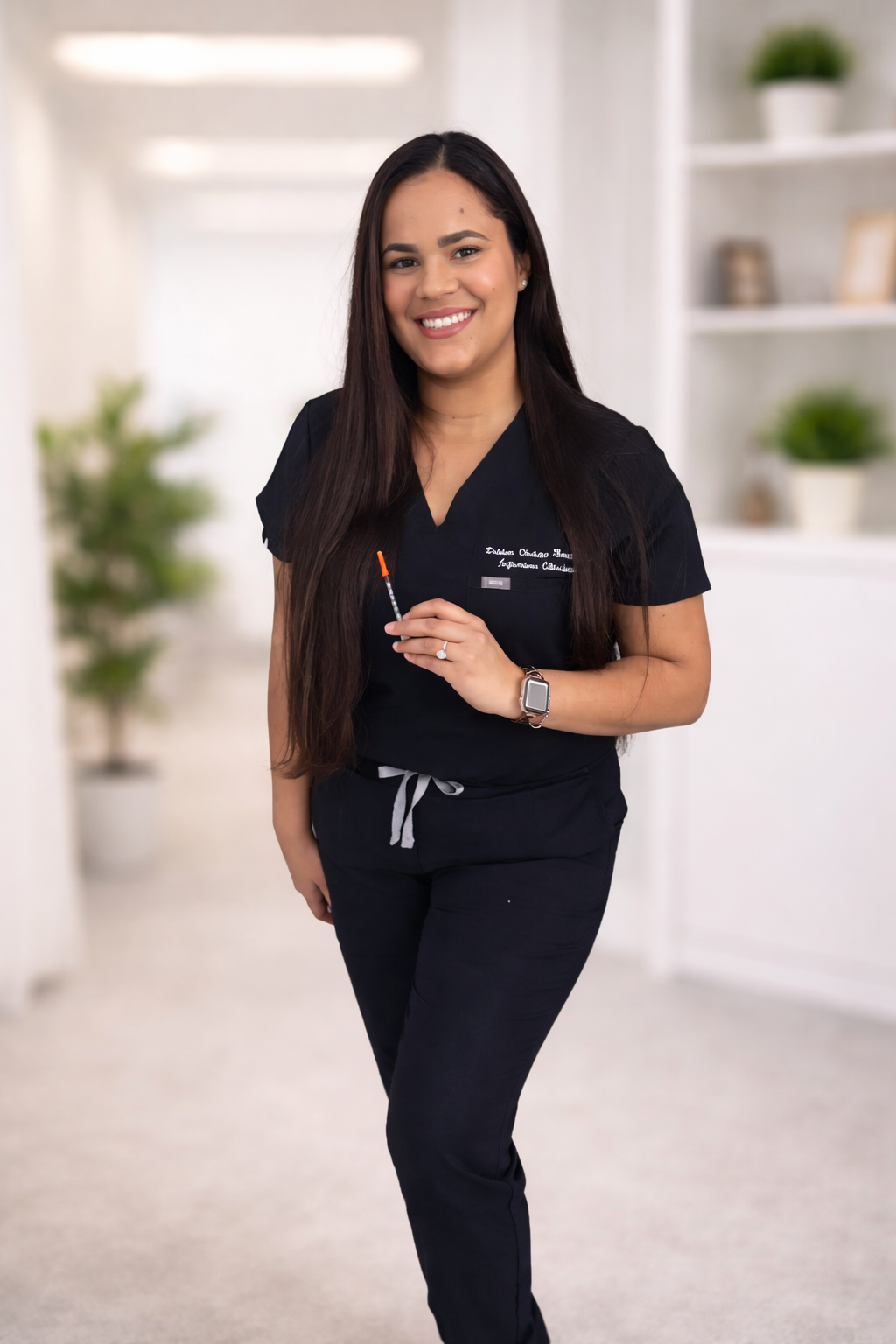 A smiling woman in medical scrubs holding a hypodermic needle, standing in a bright room with white walls, plants, and shelves.